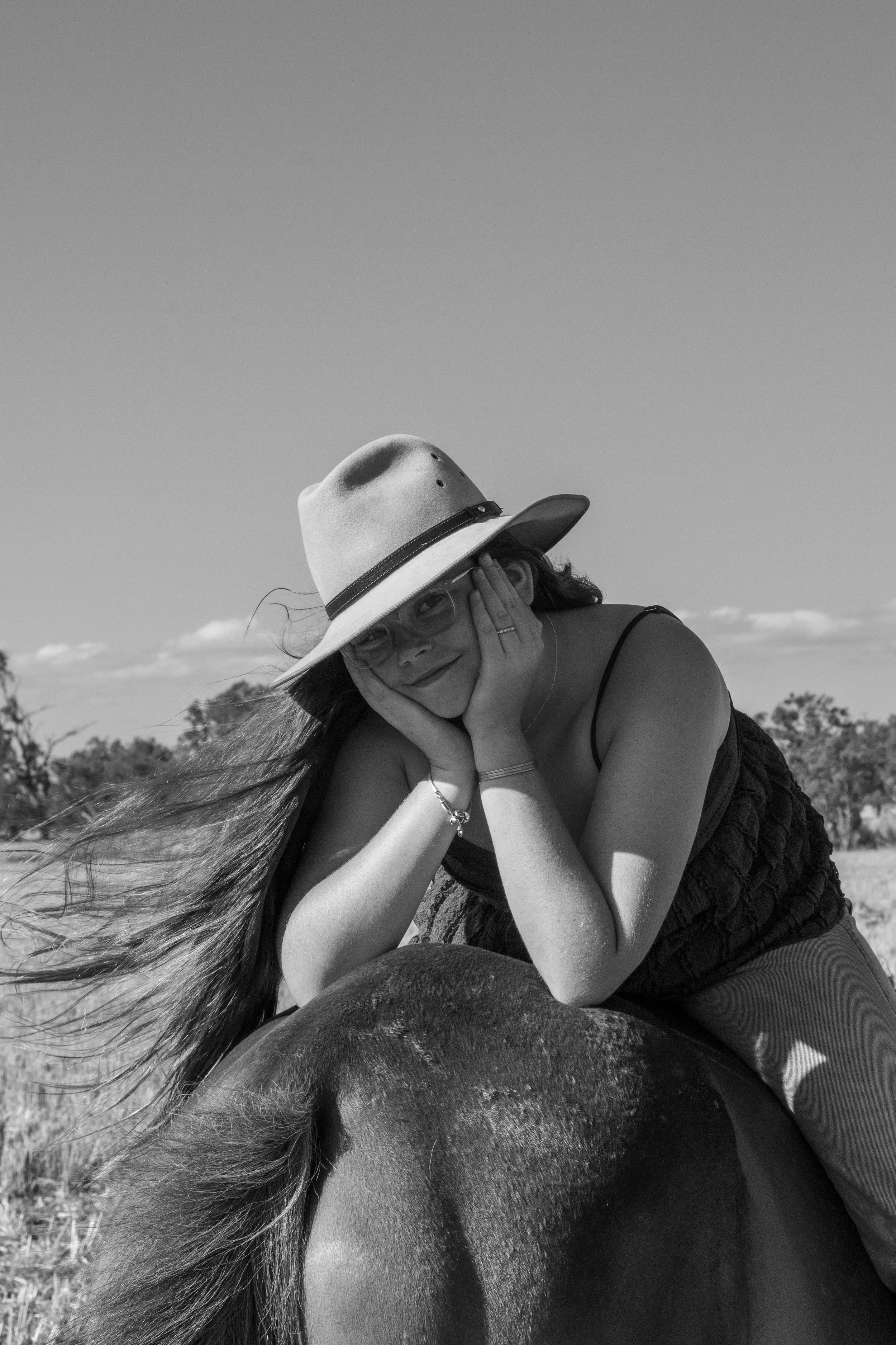 A woman in a hat and glasses lying on a horse outdoors in a field, smiling with her head resting on her hands.