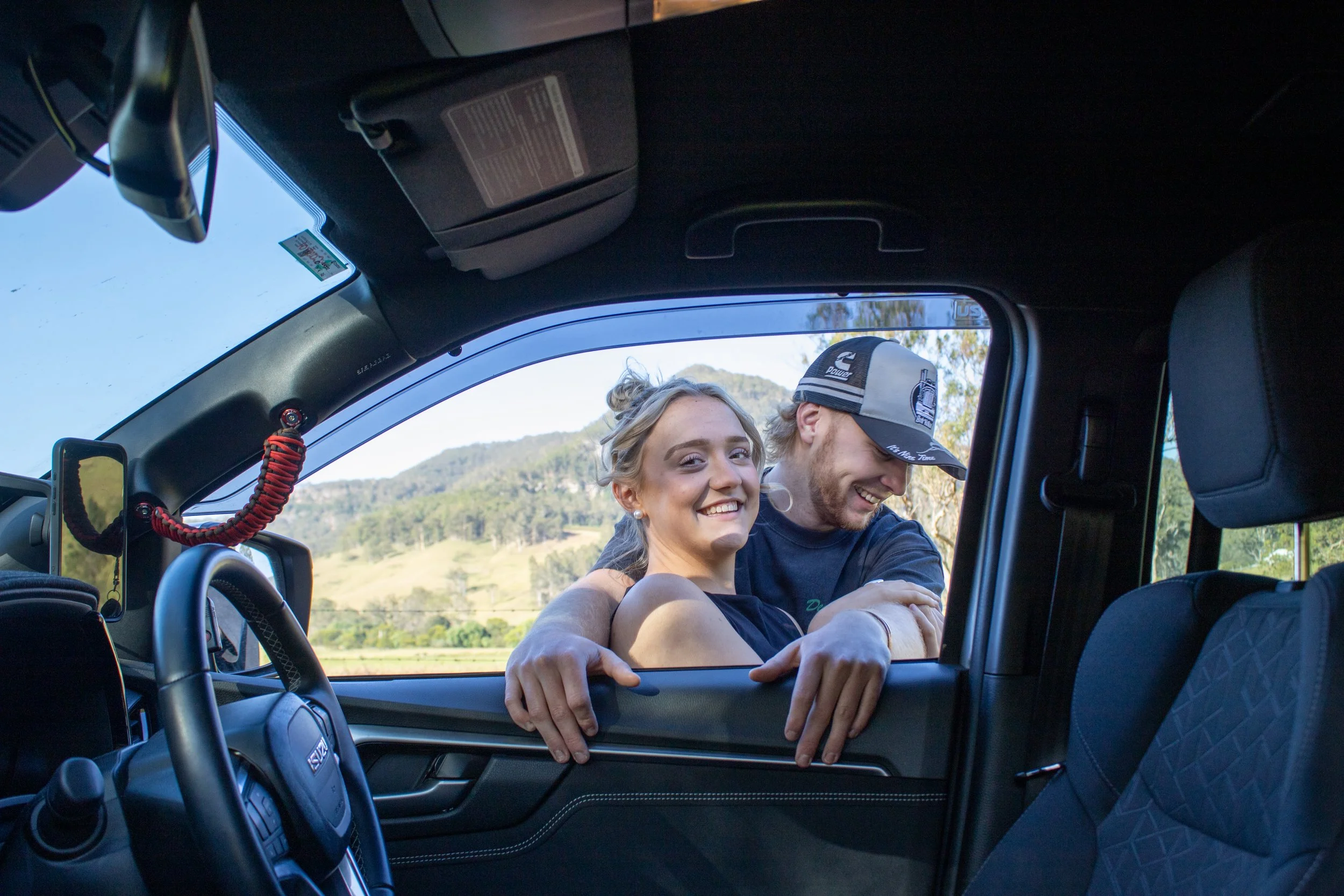 A smiling young woman and a man leaning out of a vehicle's window, happy and embracing outdoors with a scenic mountainous landscape in the background.
