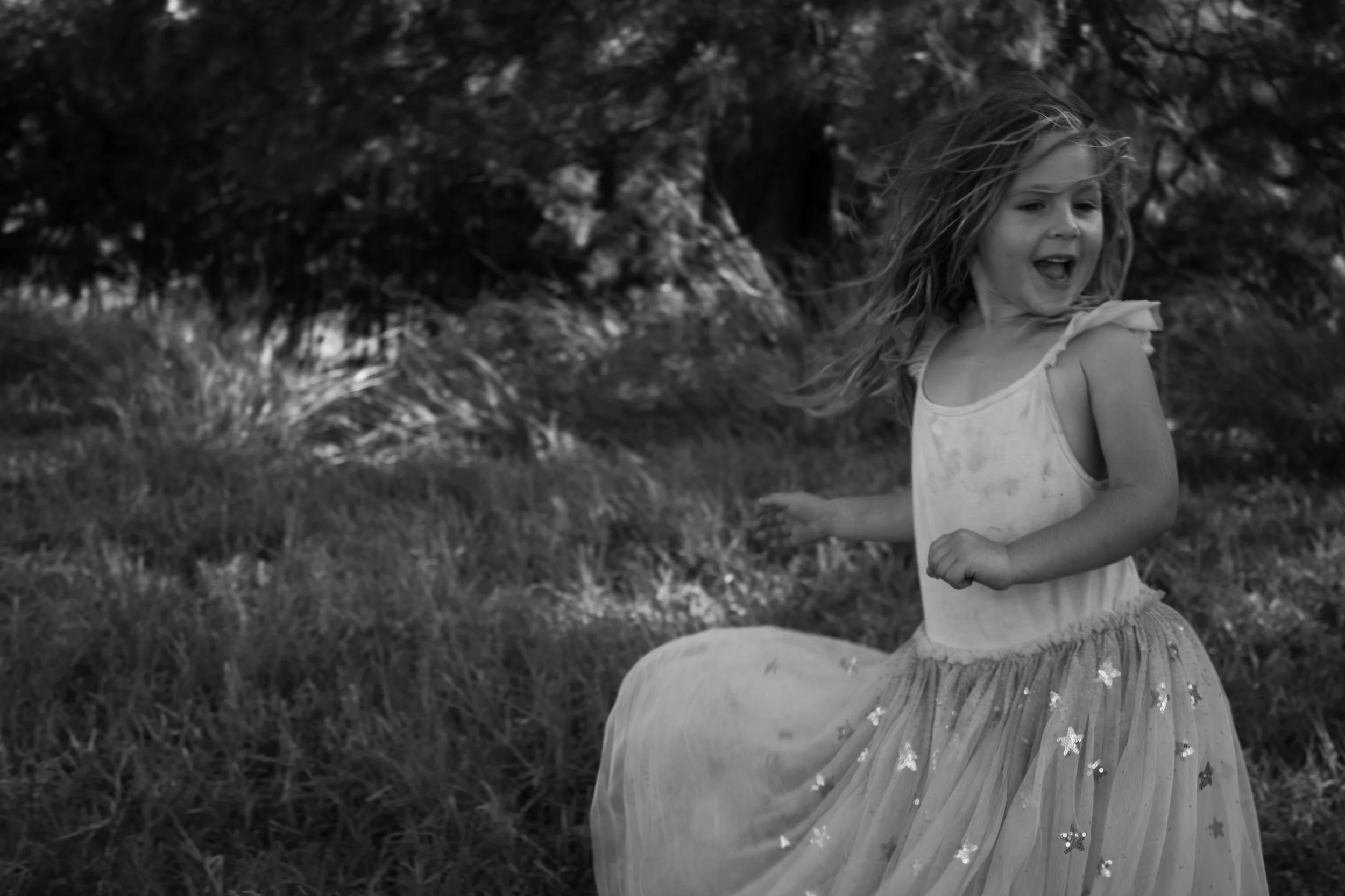 A young girl with long hair running or playing outdoors near trees, wearing a sleeveless top and a sparkly tulle skirt.