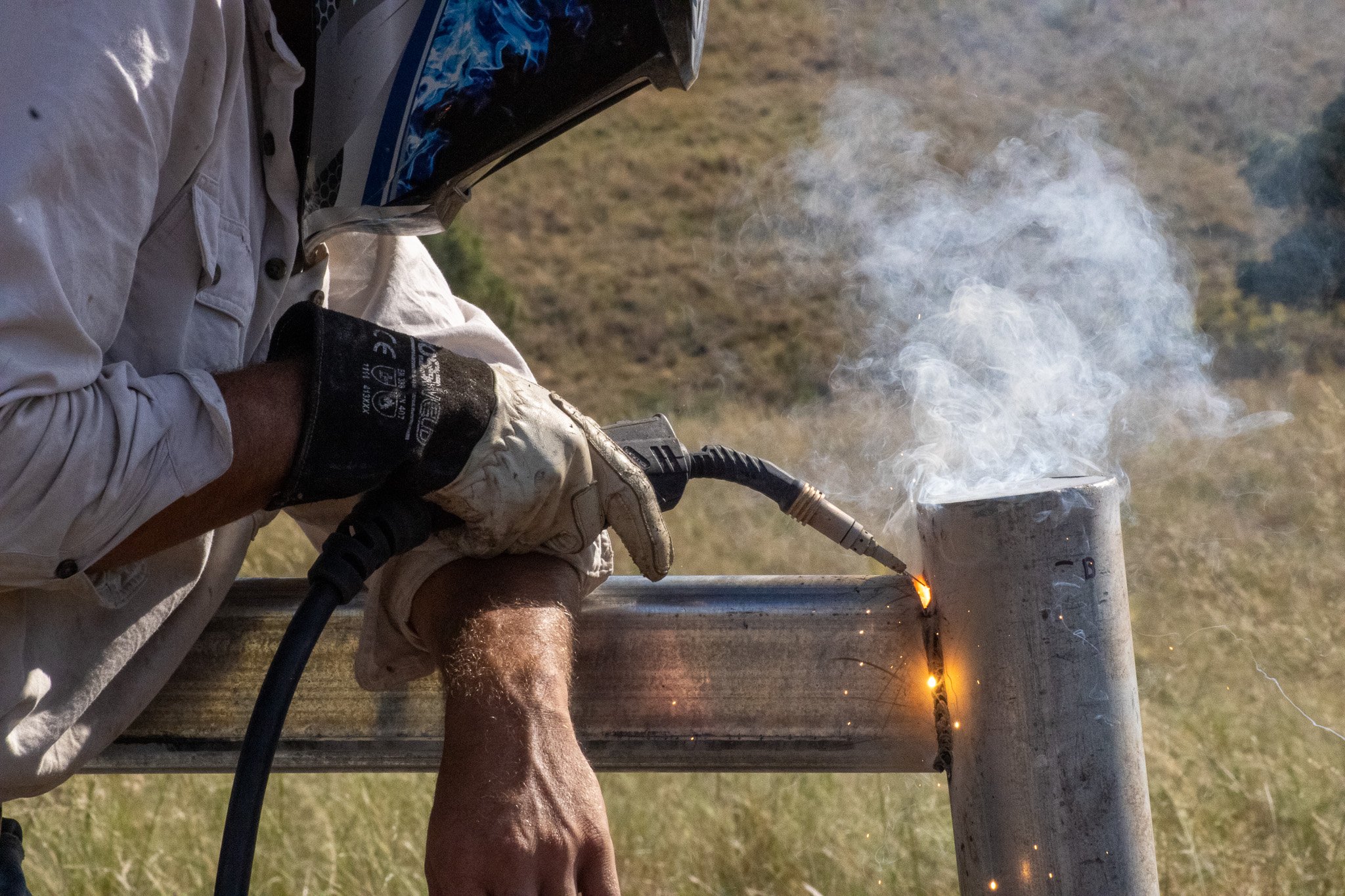 A person welding a metal pipe outdoors, wearing a welding helmet and gloves, with sparks and smoke visible.