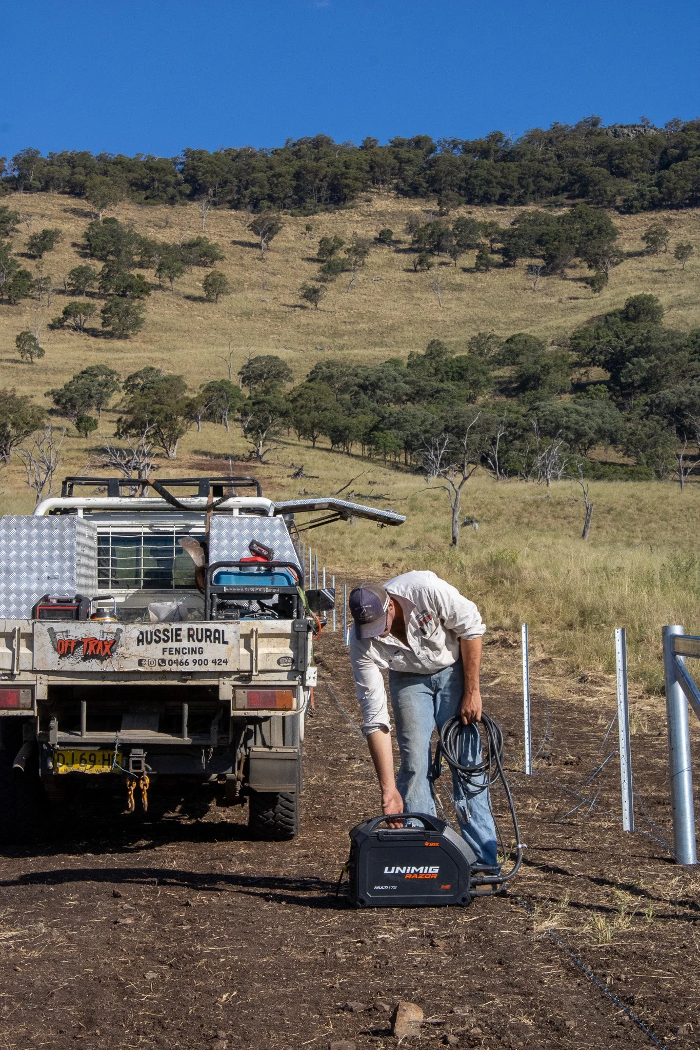 A man working on fencing in a rural, grassy landscape with hills and scattered trees, standing next to a truck with fencing equipment, using a power tool.