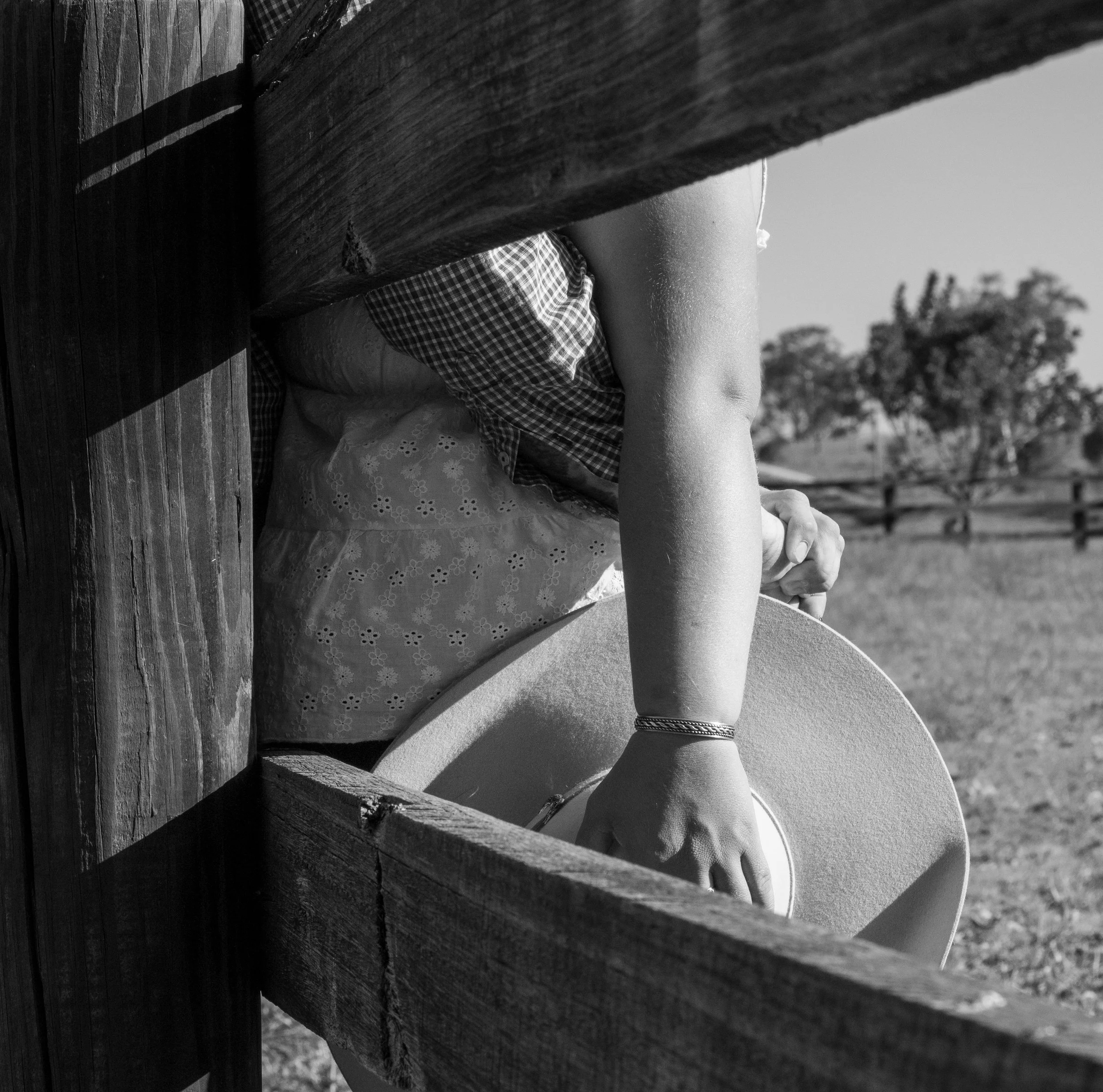 A person sitting outdoors, holding a hat, with trees and a fence in the background, viewed through a wooden fence or structure.