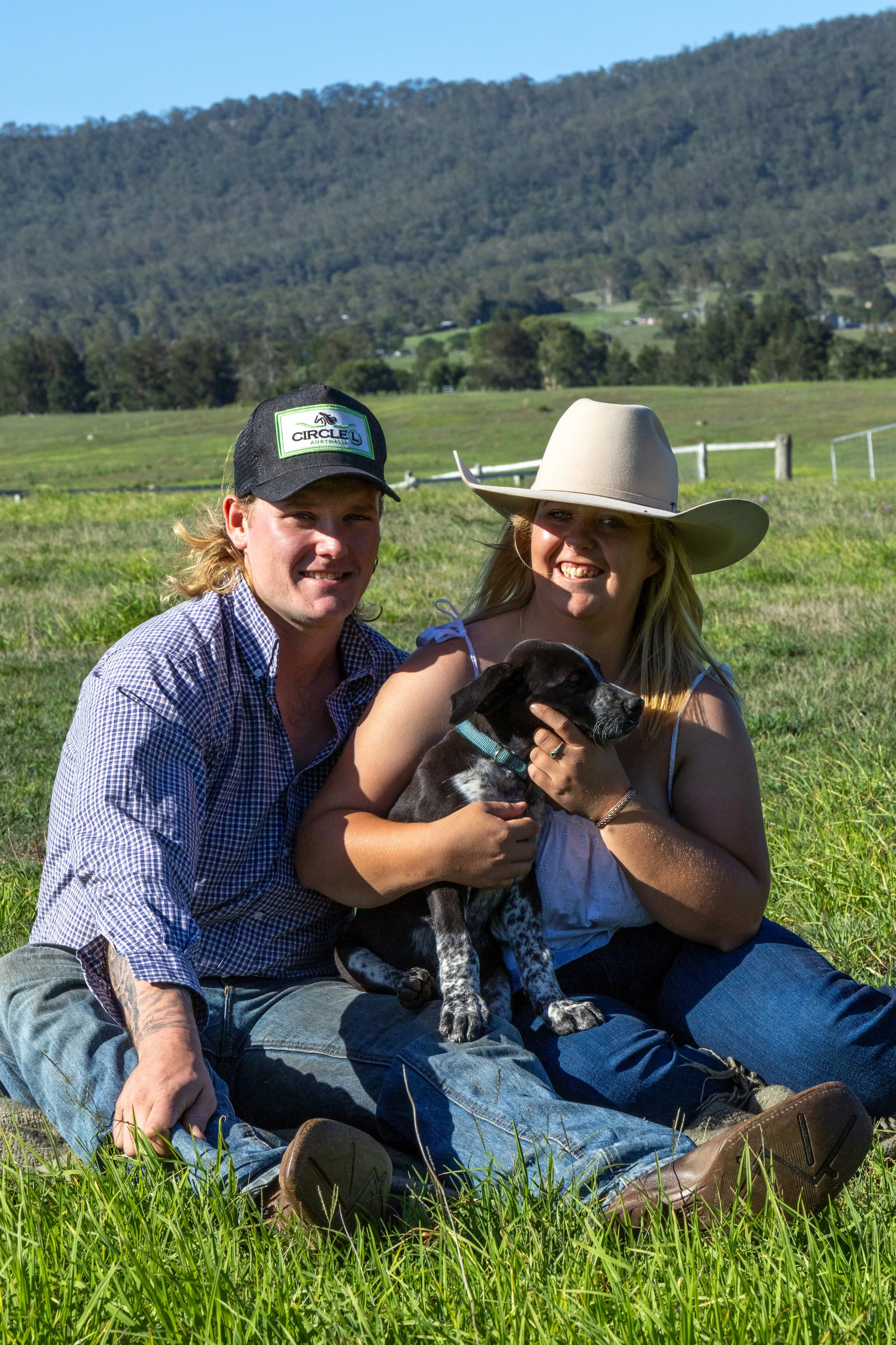 A couple sitting on grass in a field, smiling, with a woman in a large cowboy hat holding a black and white puppy, with green pasture, fences, and hills in the background.