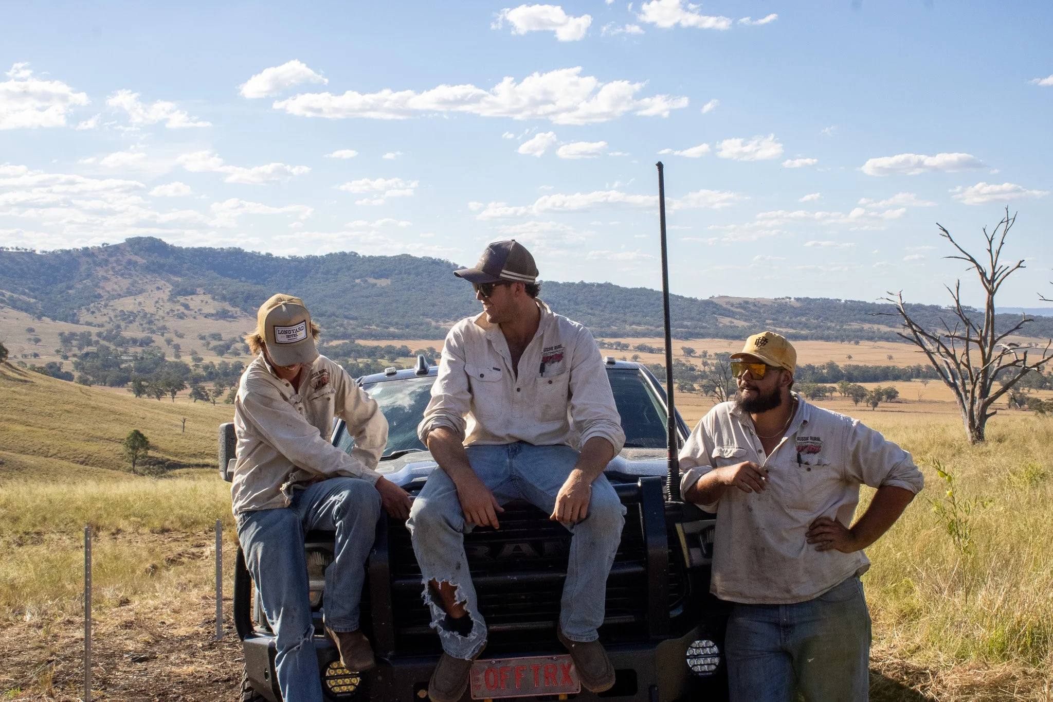 Three men in casual outdoor work clothes, one sitting on the front of a black off-road vehicle and two standing beside it, in a grassy, hilly landscape with a few trees, under a partly cloudy sky.