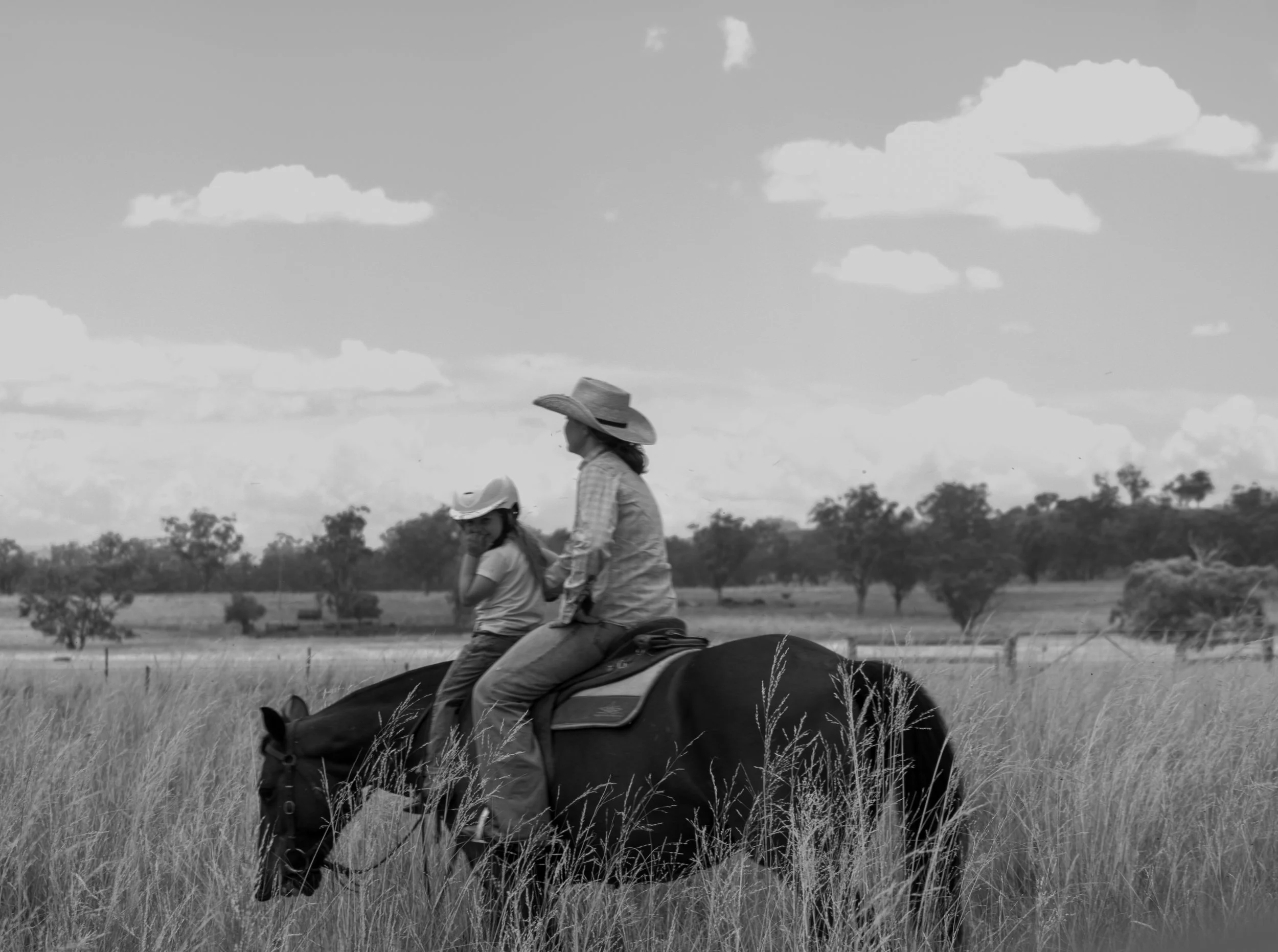 A woman and a girl riding a horse through a field of tall grass, with trees and a cloudy sky in the background.