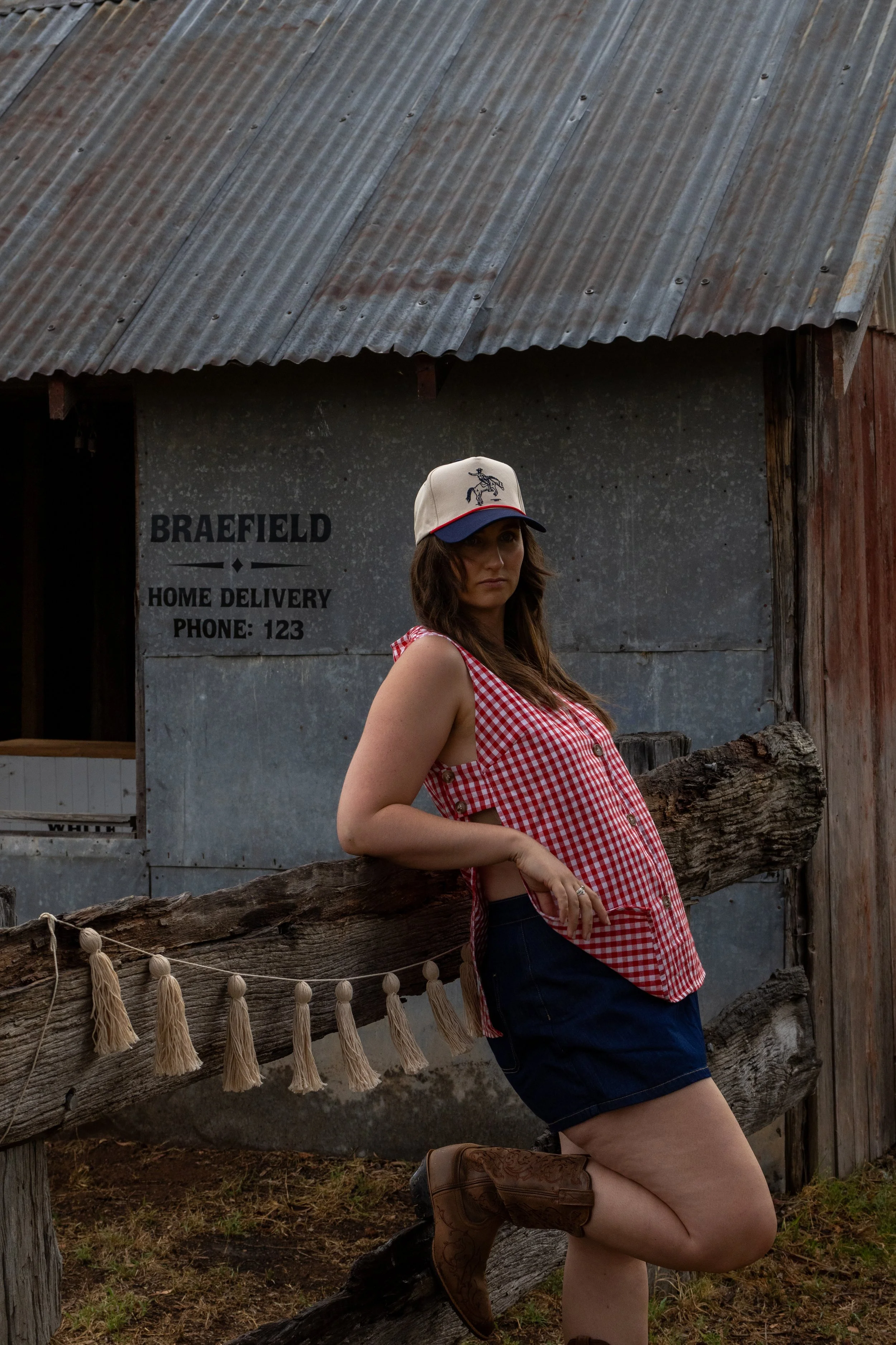 Woman in cowboy hat, red gingham sleeveless shirt, and denim shorts leaning against wooden fence in rural setting.