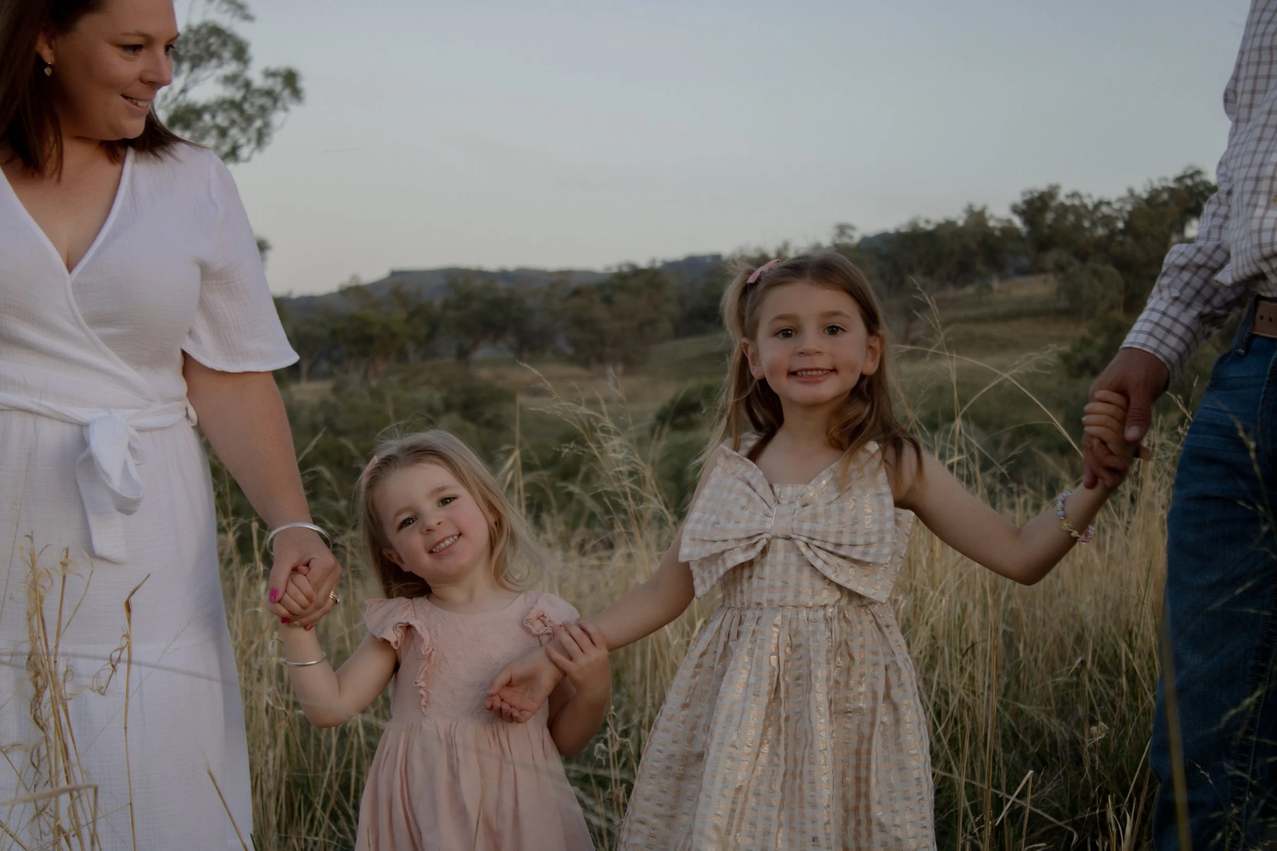 A family of four holding hands outdoors in a grassy field, with two young girls and their parents, smiling at the camera during the evening.