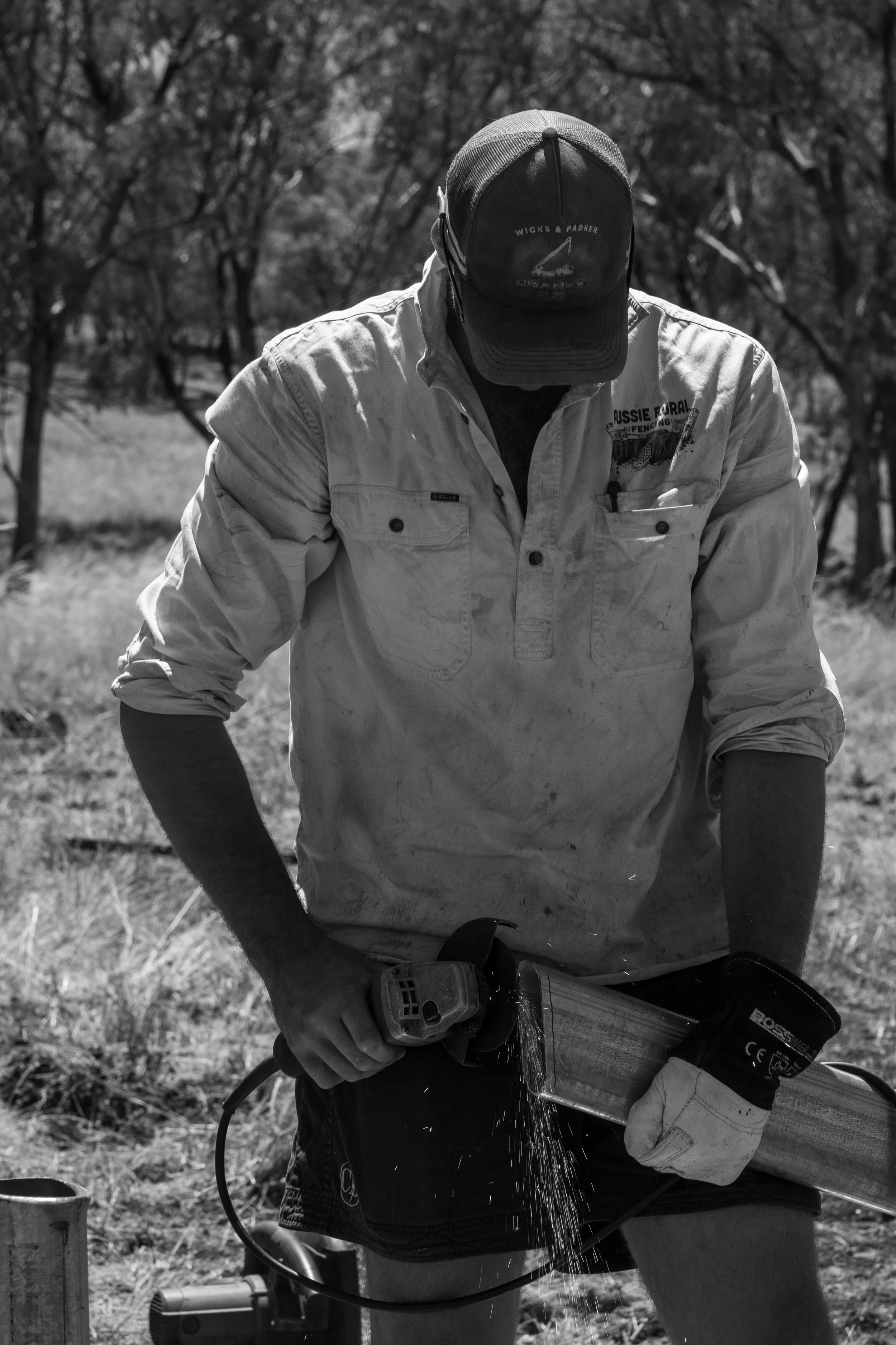 A man wearing a cap, long-sleeved shirt, and gloves is cutting a metal pipe outdoors with an angle grinder, with sparks flying.