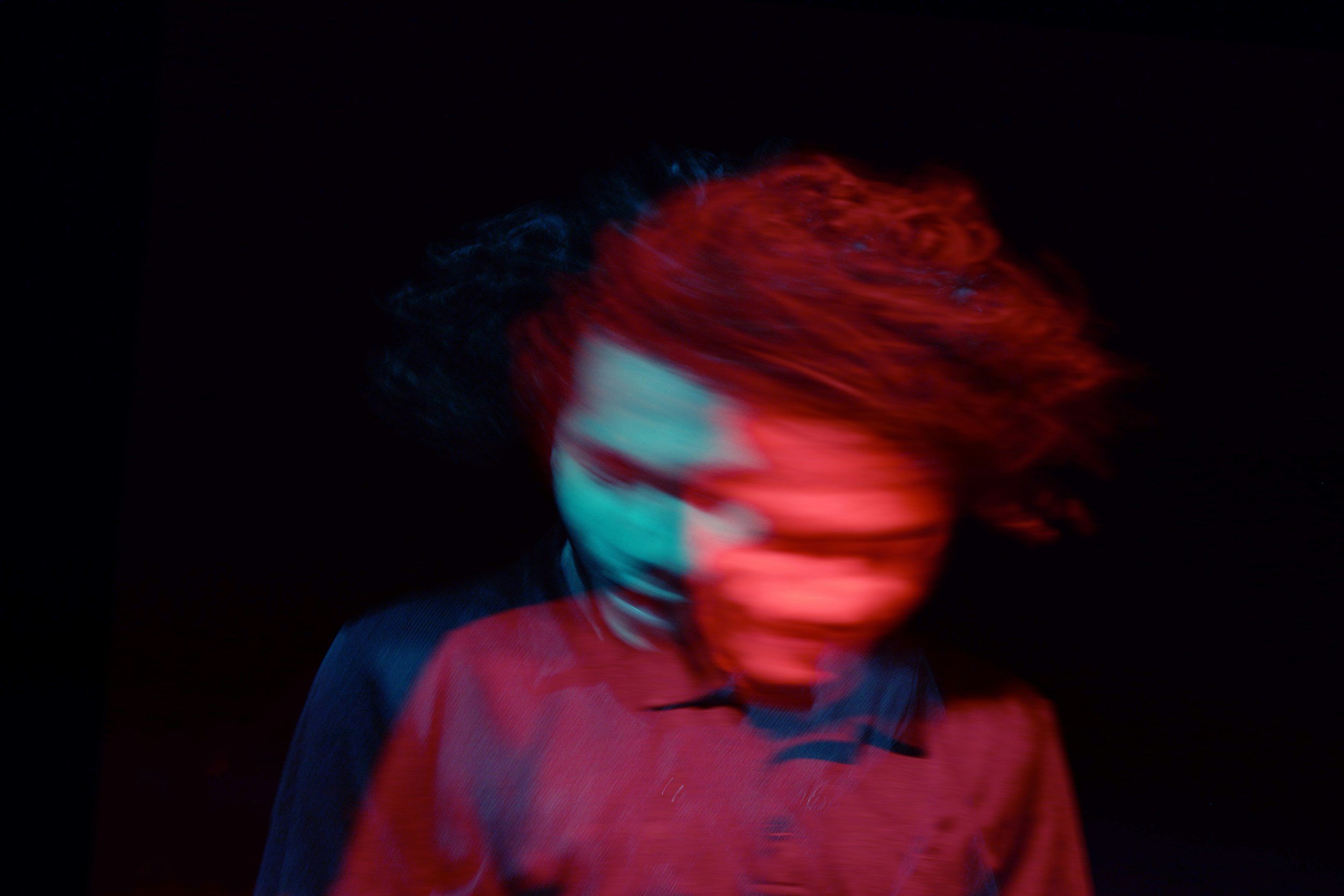 A person with curly hair moving quickly, creating a blurred motion effect with red, blue, and black tones against a dark background.