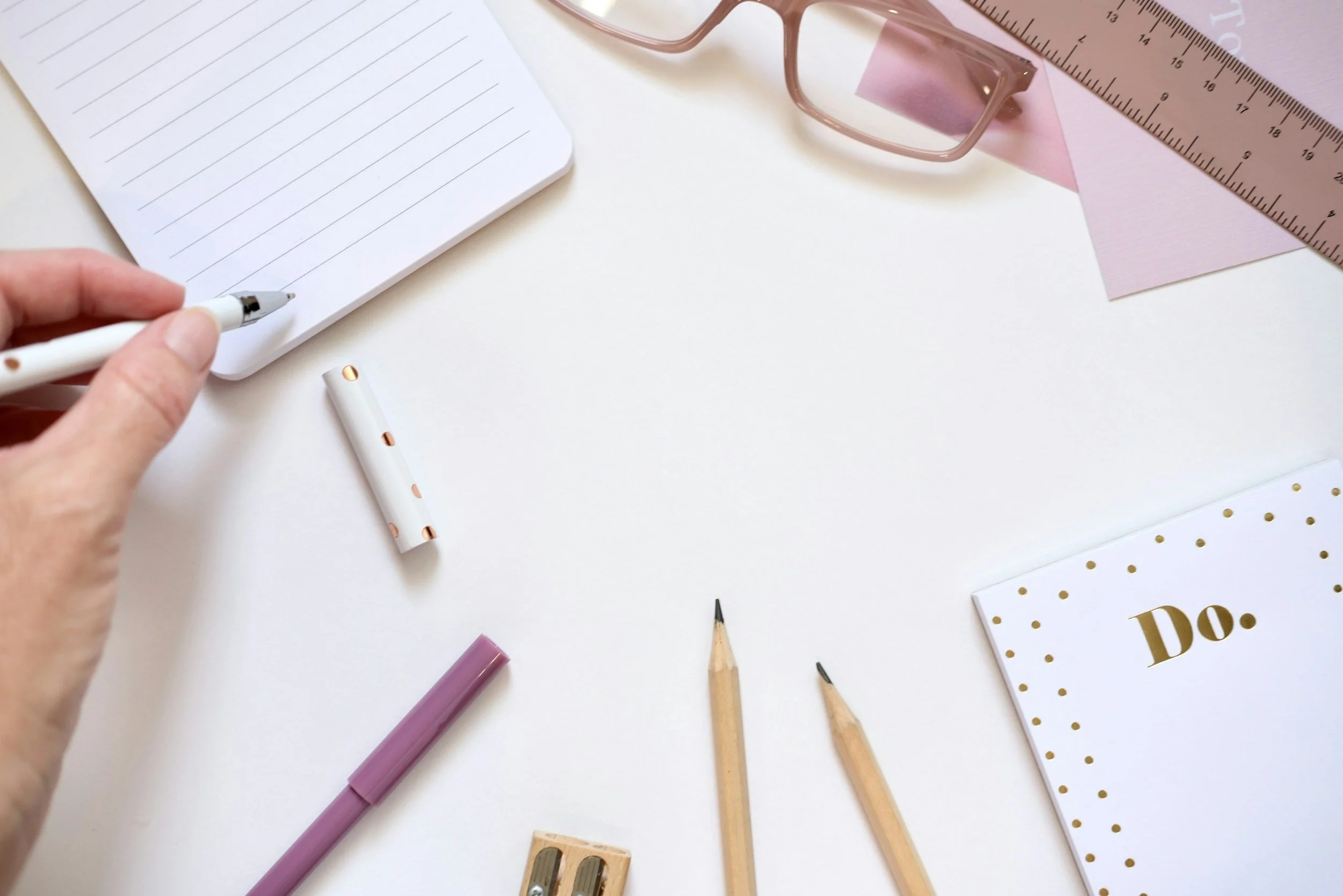 Flat lay of stationery items including a notepad, pens, a ruler, glasses, and a sticky note on a white surface.