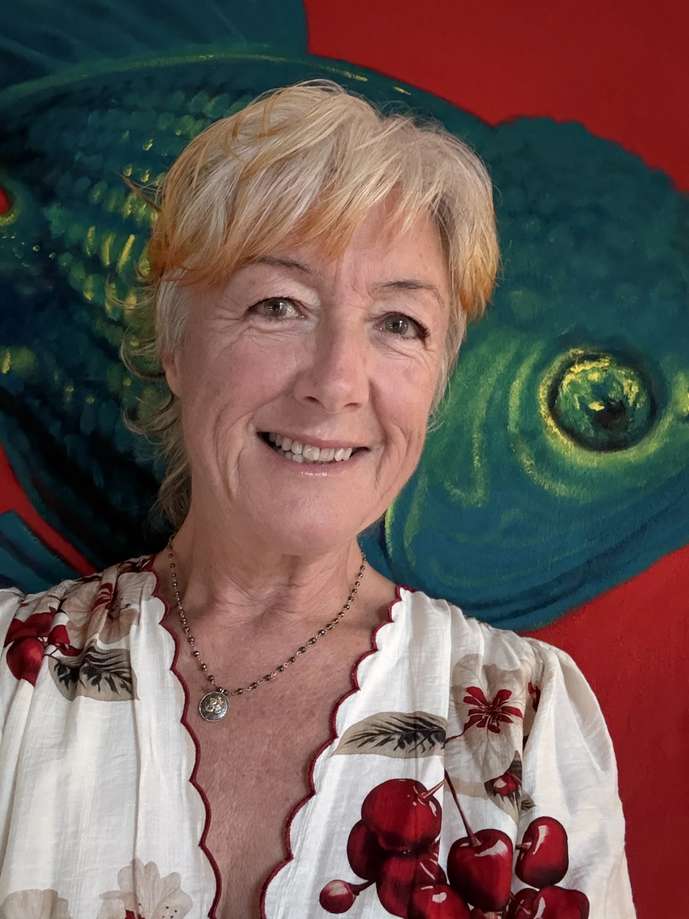 A smiling woman with short blonde hair taking a selfie in front of a colorful chameleon painting. She is wearing a white blouse with a cherry pattern and a silver necklace.