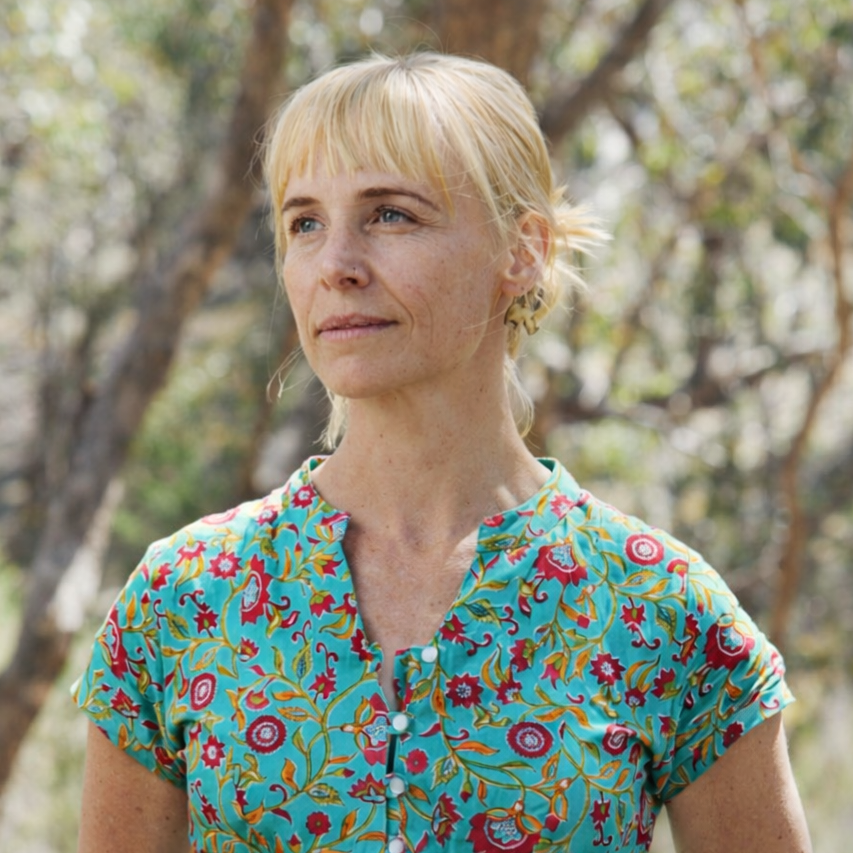 A woman with blonde hair and a nose piercing looking off-camera outdoors, wearing a colorful floral blouse with a nature background.