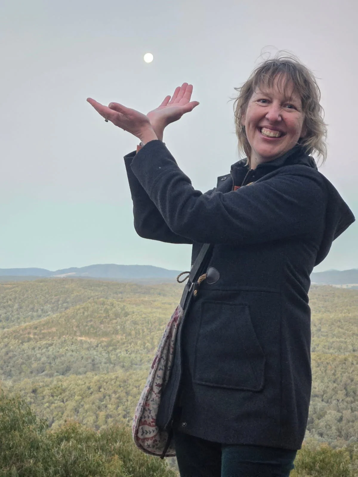 Woman with curly blonde hair smiling and gesturing towards the sky with her hands, standing outdoors against a backdrop of rolling green hills and a cloudy sky with the moon visible.
