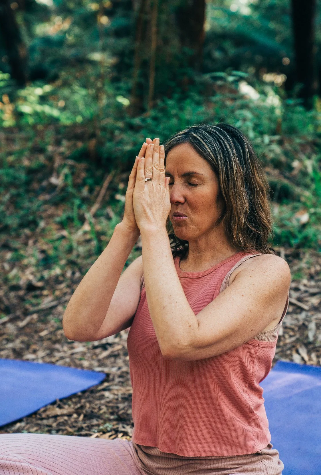 A woman practicing yoga outdoors in a wooded area, sitting cross-legged with palms pressed together near her face, eyes closed, wearing a sleeveless coral top and pink pants.