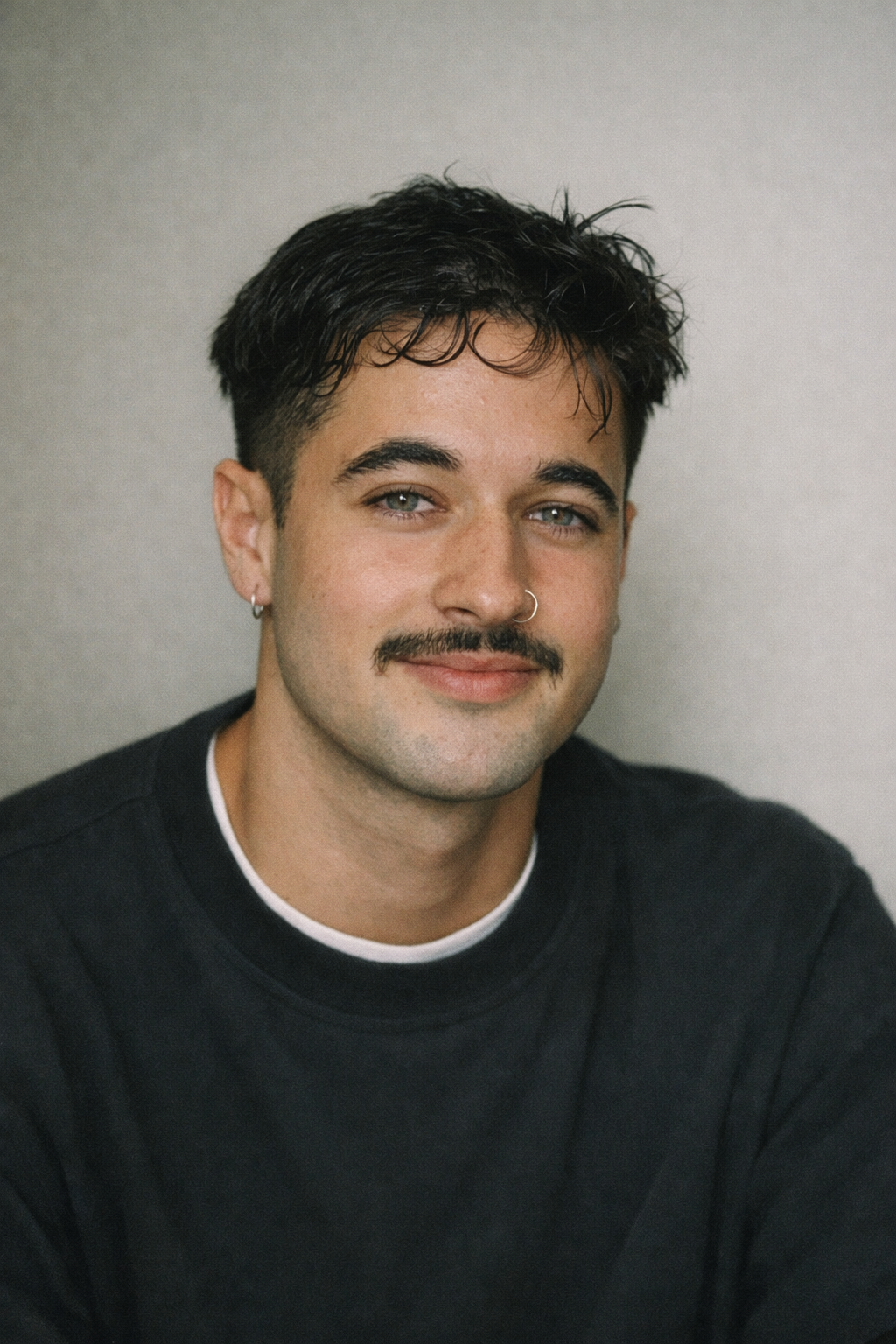 A young man with dark, curly hair, a mustache, and a nose ring, smiling at the camera against a plain background.