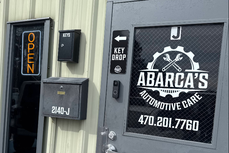 Outside view of Arabaca's Automotive Care shop with an illuminated 'OPEN' sign, a key drop box, a keypad entry system, and a window with the shop's logo and contact number.