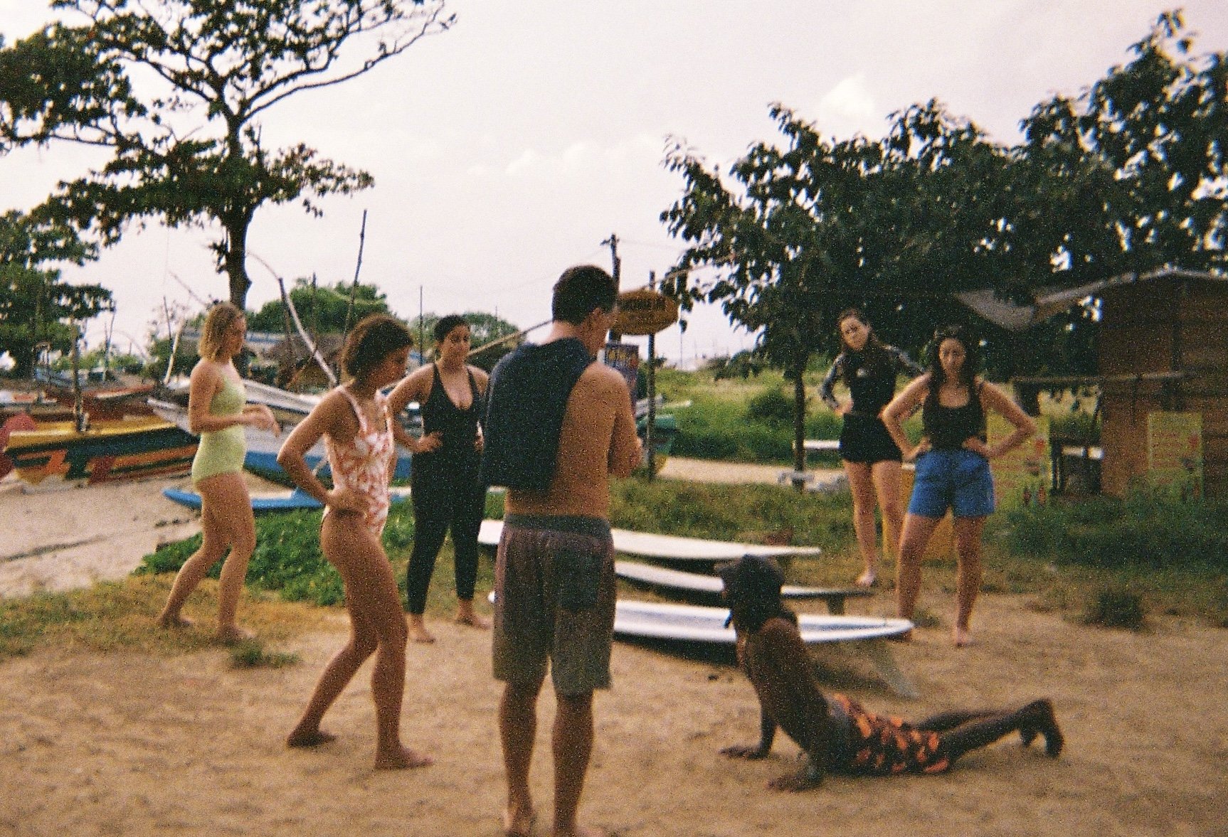 A group of people on a beach near kayaks and paddleboards, with some standing and one lying on the sand, under trees with overcast sky.