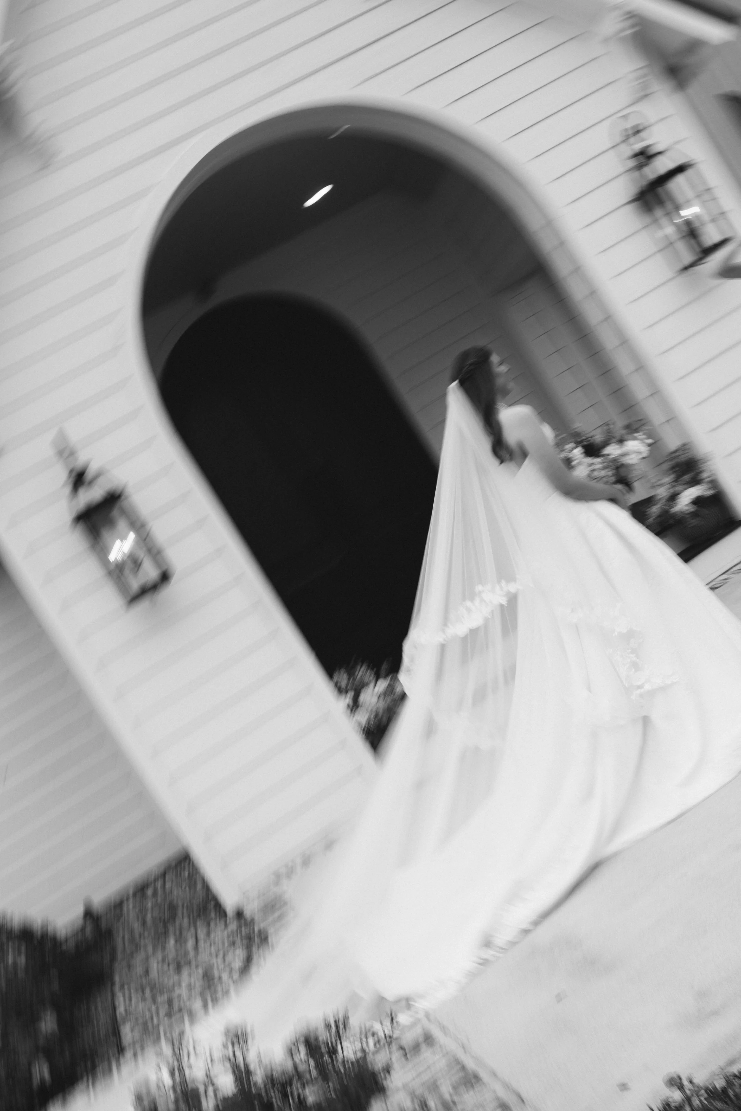 A blurry black and white image of a woman in a white dress with a veil, possibly a bride, standing outside a building with an archway and lanterns on the wall.