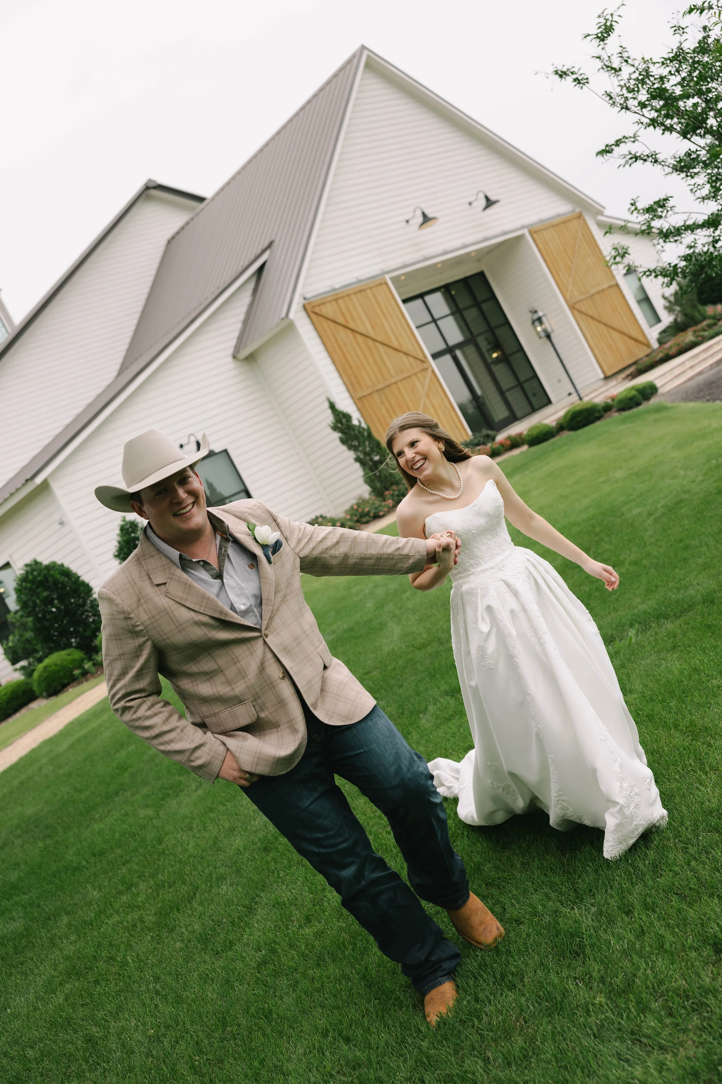 A happy bride and groom holding hands and smiling outside a modern white house with wooden accents, green lawn, and overcast sky.