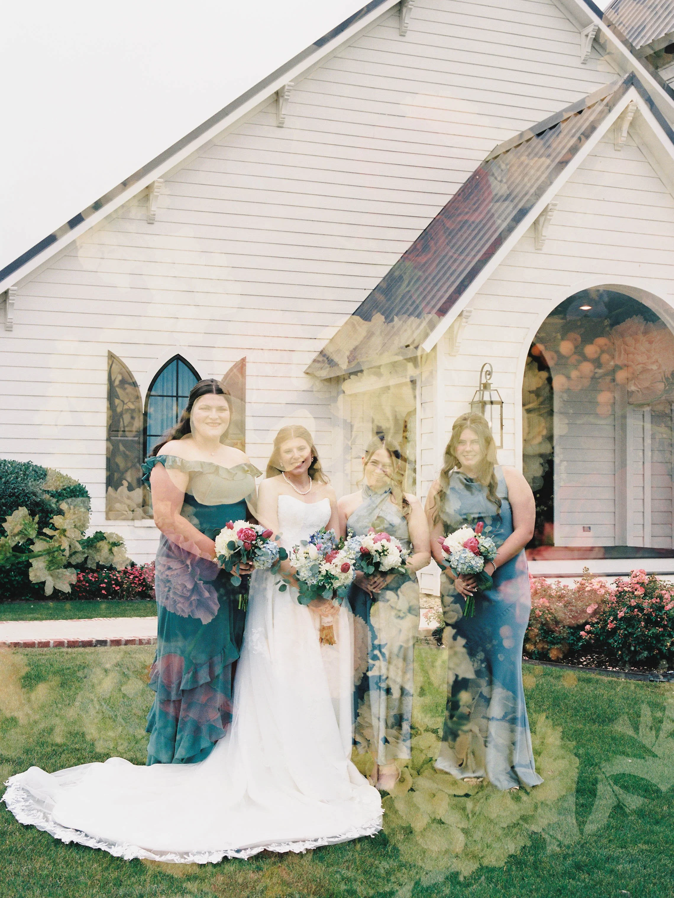 A group of women dressed in formal attire holding bouquets of flowers, standing in front of a small white church with a rainbow stained glass window and pink flowers in the garden.