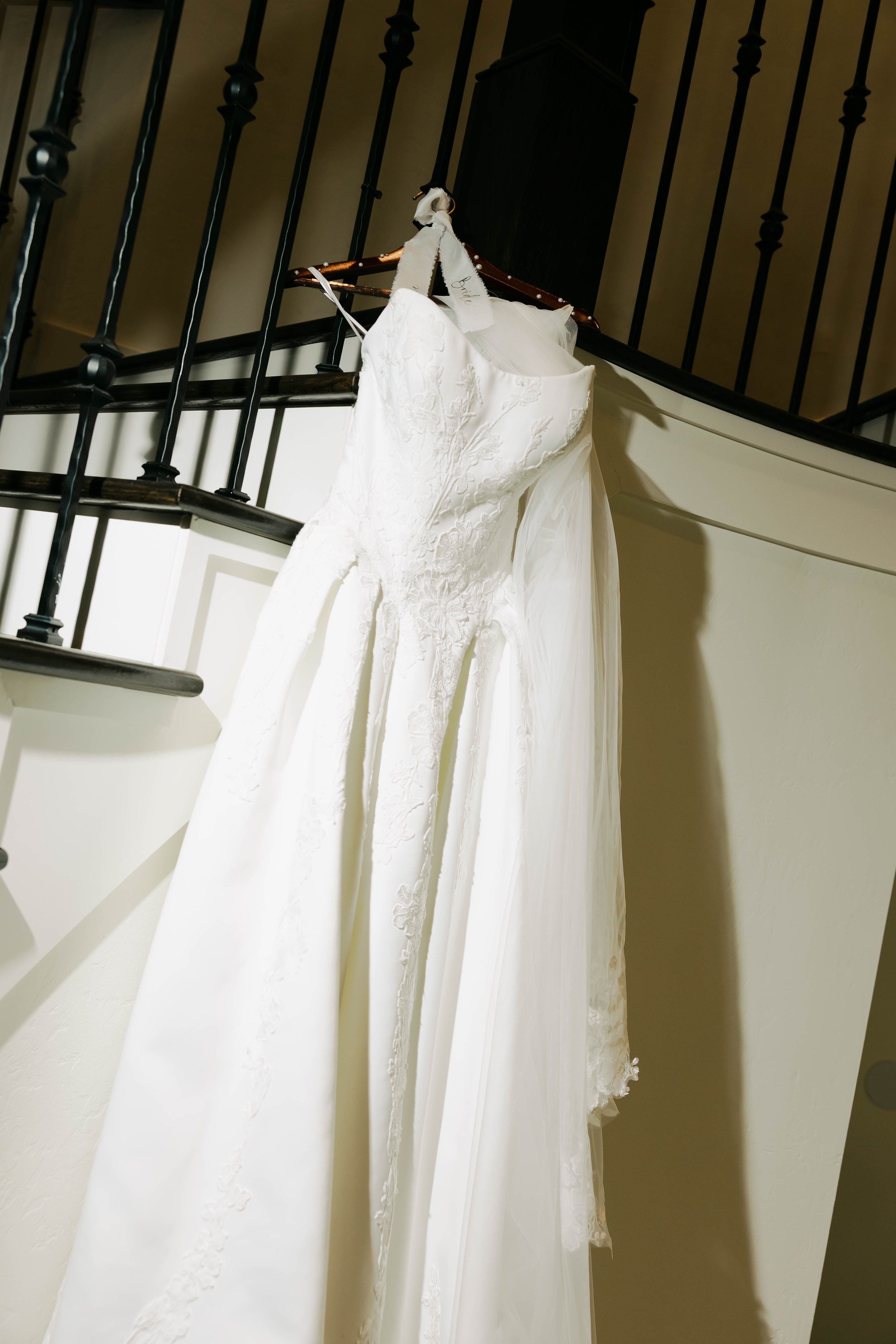 A white wedding dress with lace details hanging on a wooden hanger on a staircase railing.