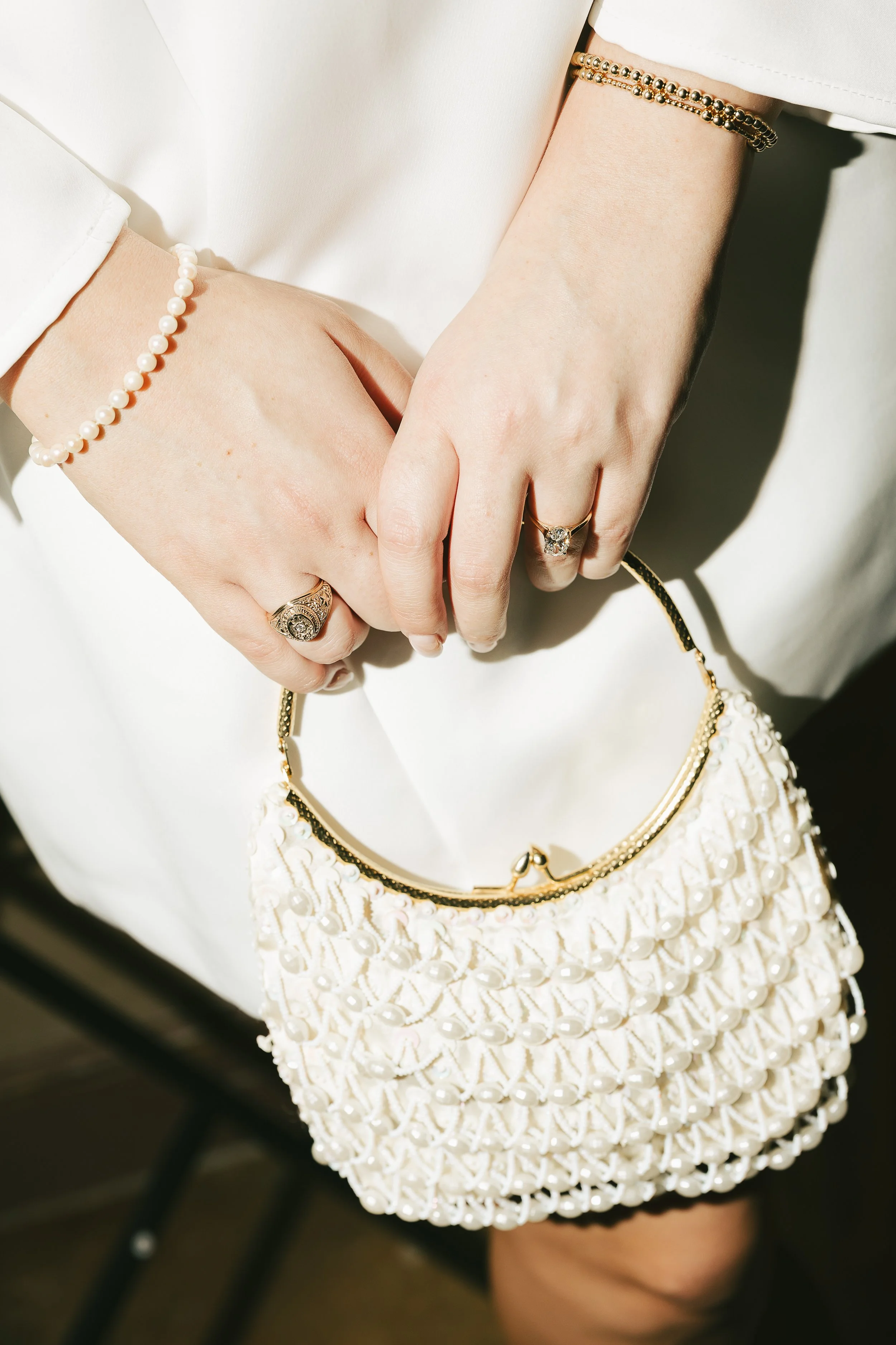 Close-up of two hands with rings, jewelry, and a pearl-embellished purse, holding onto a white handbag.