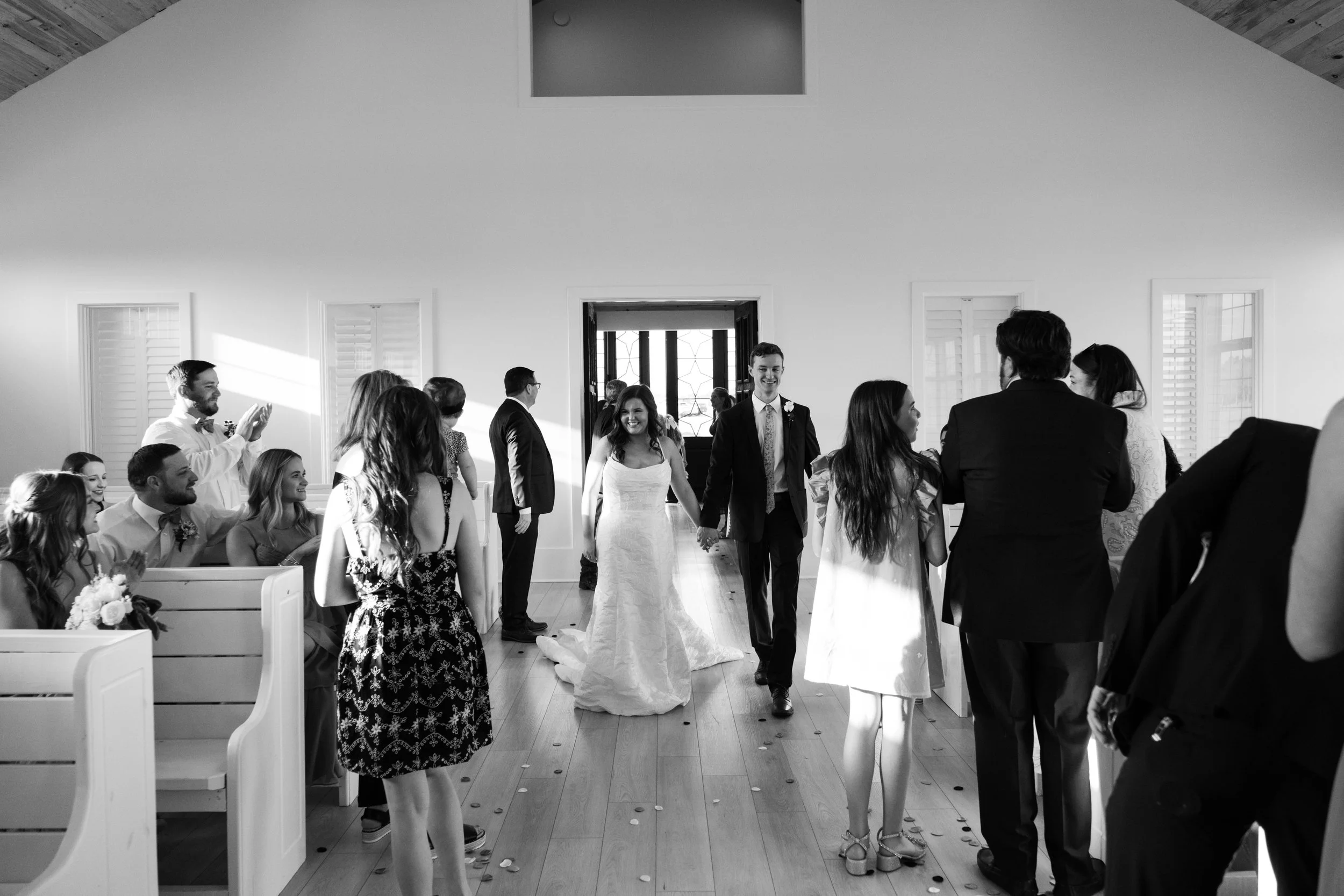 A wedding ceremony with the bride and groom holding hands and walking down the aisle in a bright, modern church. Guests are seated and standing, clapping and smiling, capturing the joyful moment.