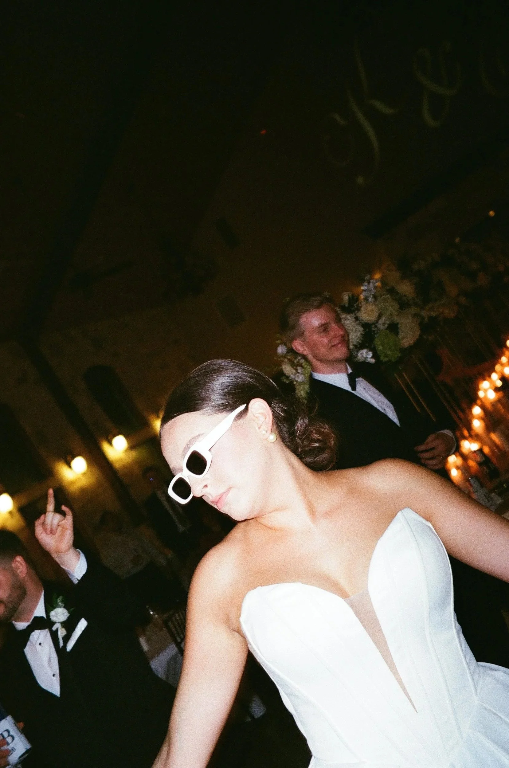 Woman in white wedding dress wearing white sunglasses, dancing at wedding reception with a man in tuxedo in the background.