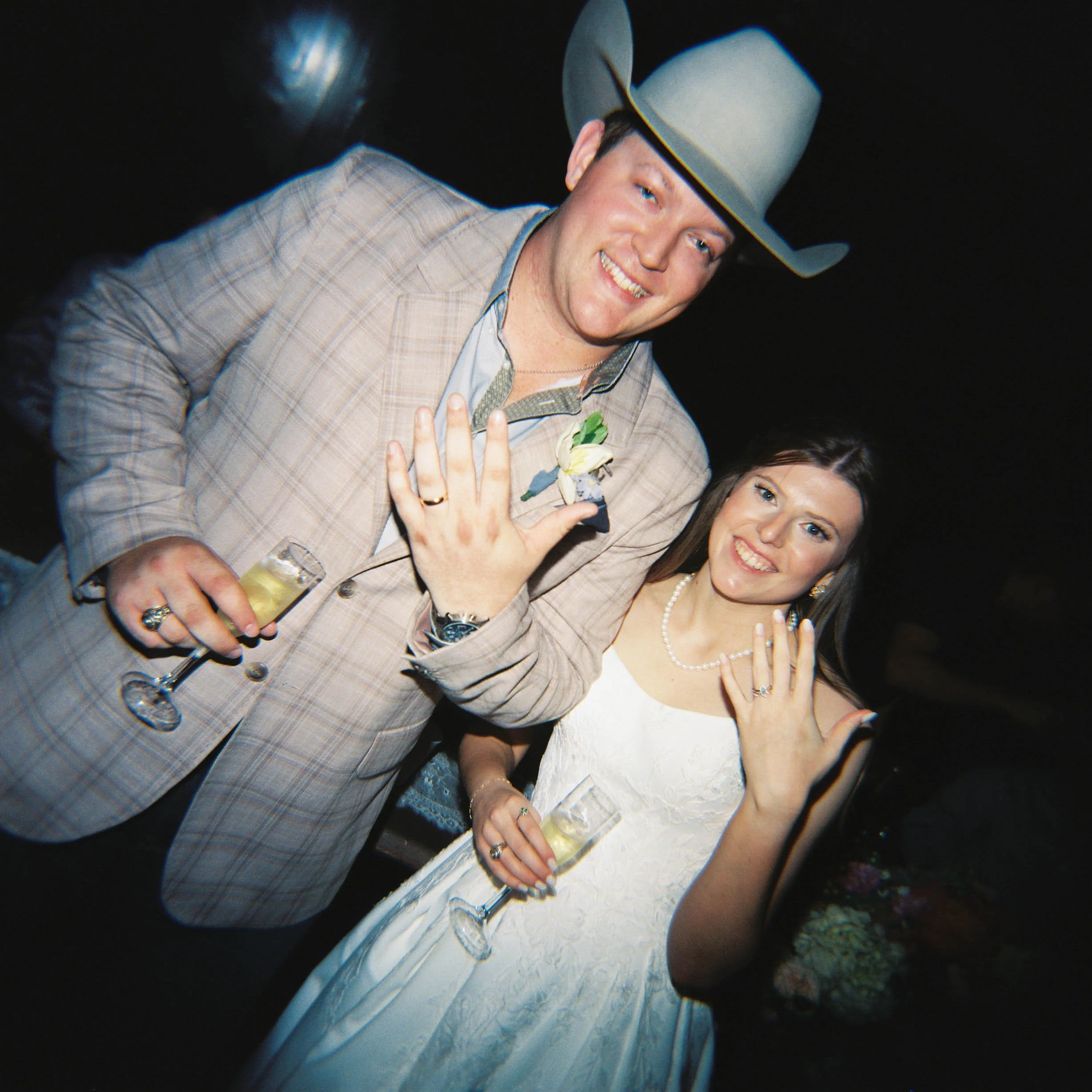 A man in a gray plaid suit and a wide-brimmed hat smiling at a celebration, holding a glass of champagne, with a woman beside him smiling and showing rings on her finger. Deep in the Heart Farms is a charming wedding venue in Brenham, Texas