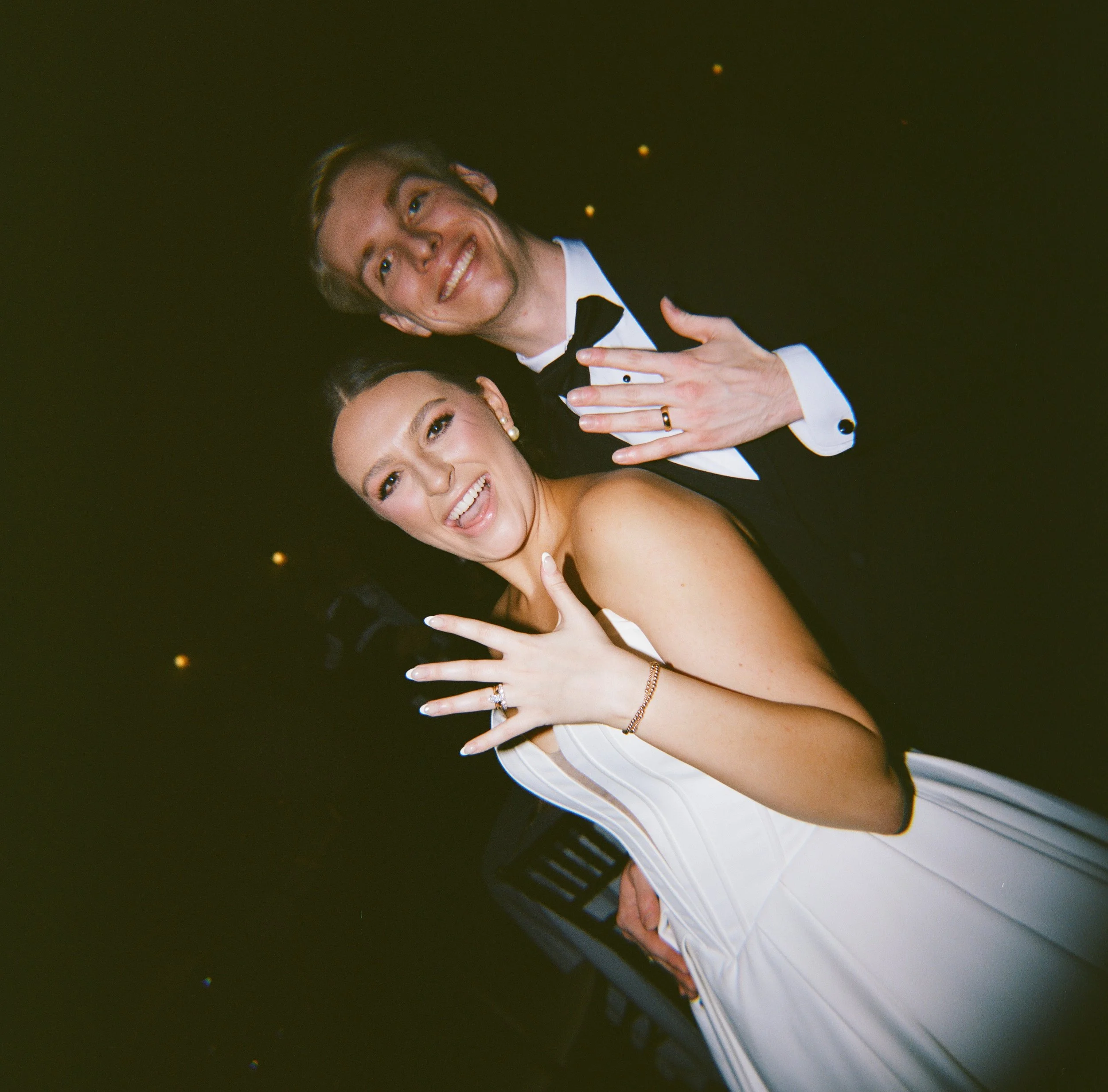 A bride and groom at a wedding reception, smiling and showing their wedding rings, with a dark background and small lights.