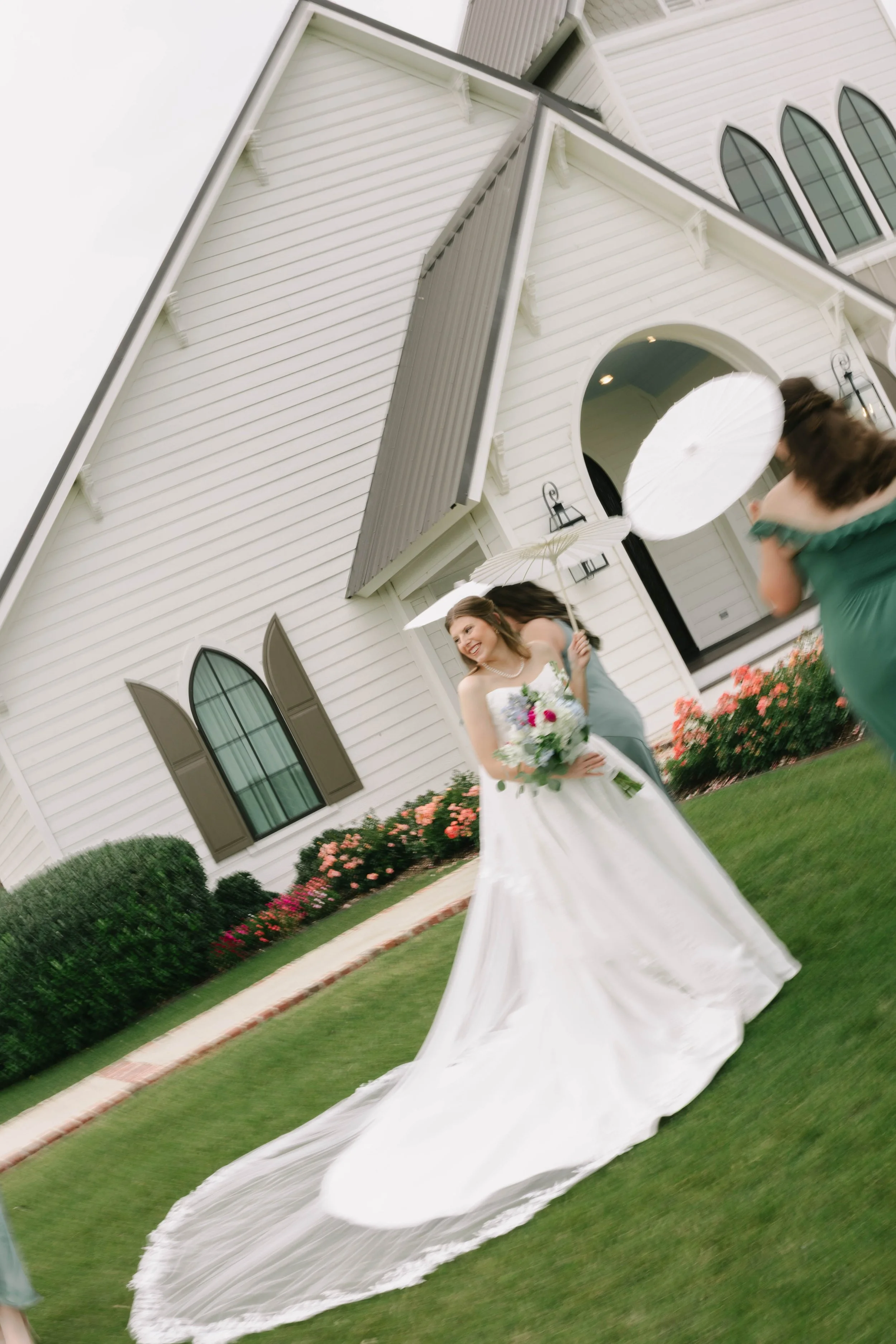 A bride in a wedding dress holding a bouquet of flowers standing on a lawn in front of a white church, with a woman and a young girl holding umbrellas nearby.