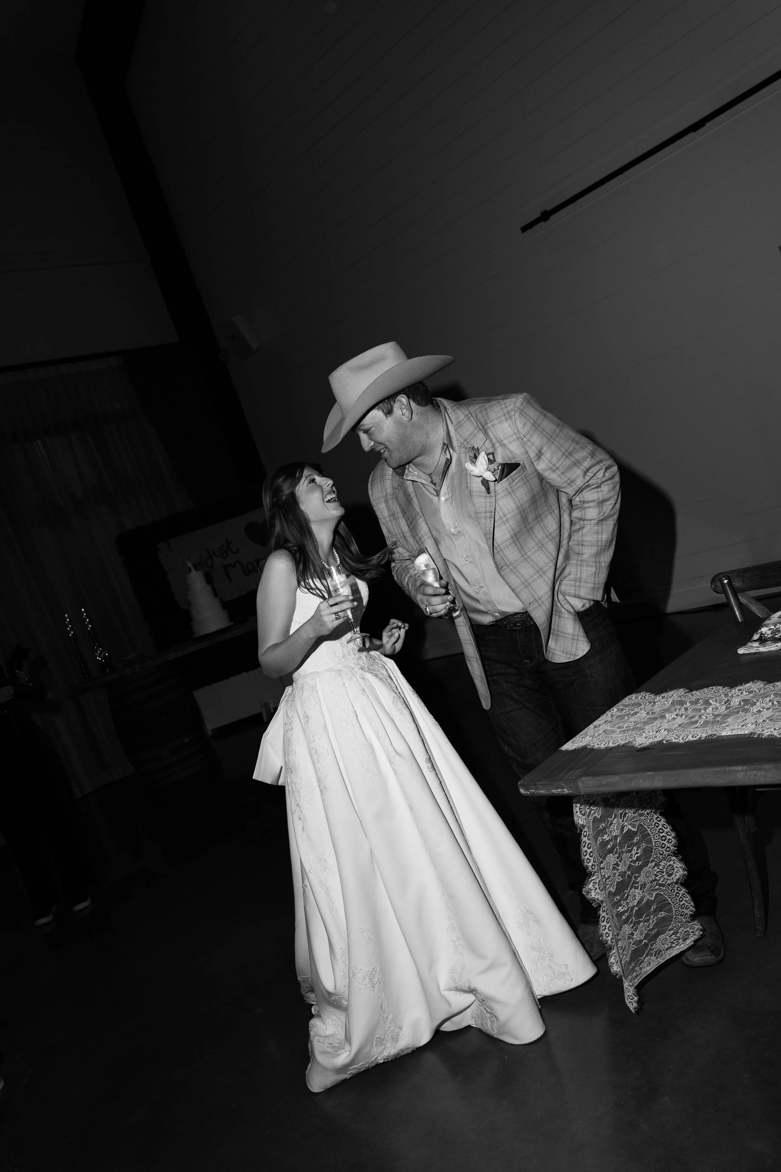 A bride and a groom sharing a moment of happiness at their wedding reception, with the bride in a white wedding dress and the groom in a plaid suit and cowboy hat.
