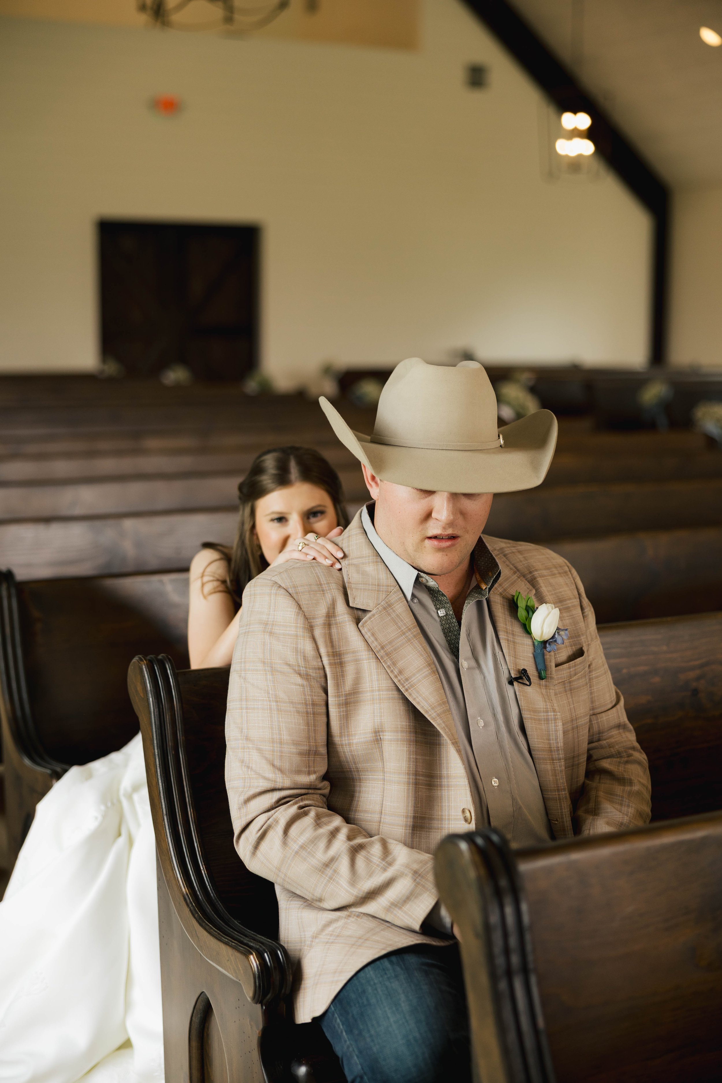 A man in a beige cowboy hat, brown plaid blazer, and blue jeans is seated in a pew with a woman in a white dress behind him. The woman is resting her hand on his shoulder and looking at him. The background shows a dimly lit interior with wooden furni