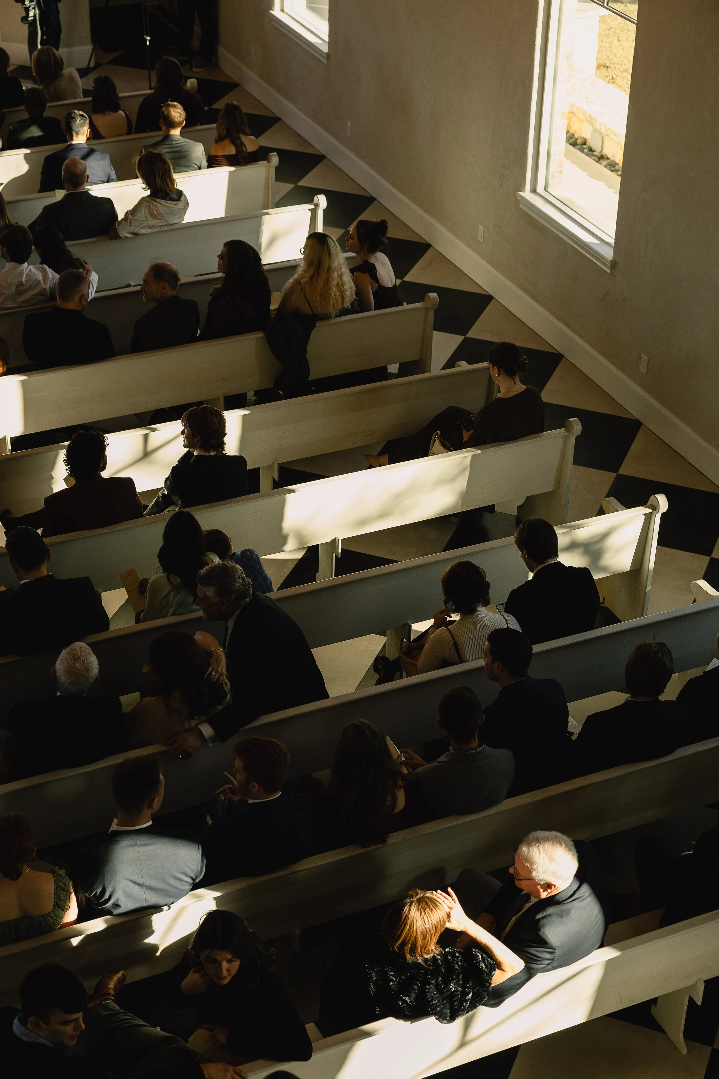 People sitting in pews inside a church during a service, sunlight coming through large windows, and some attendees engaging in conversations.