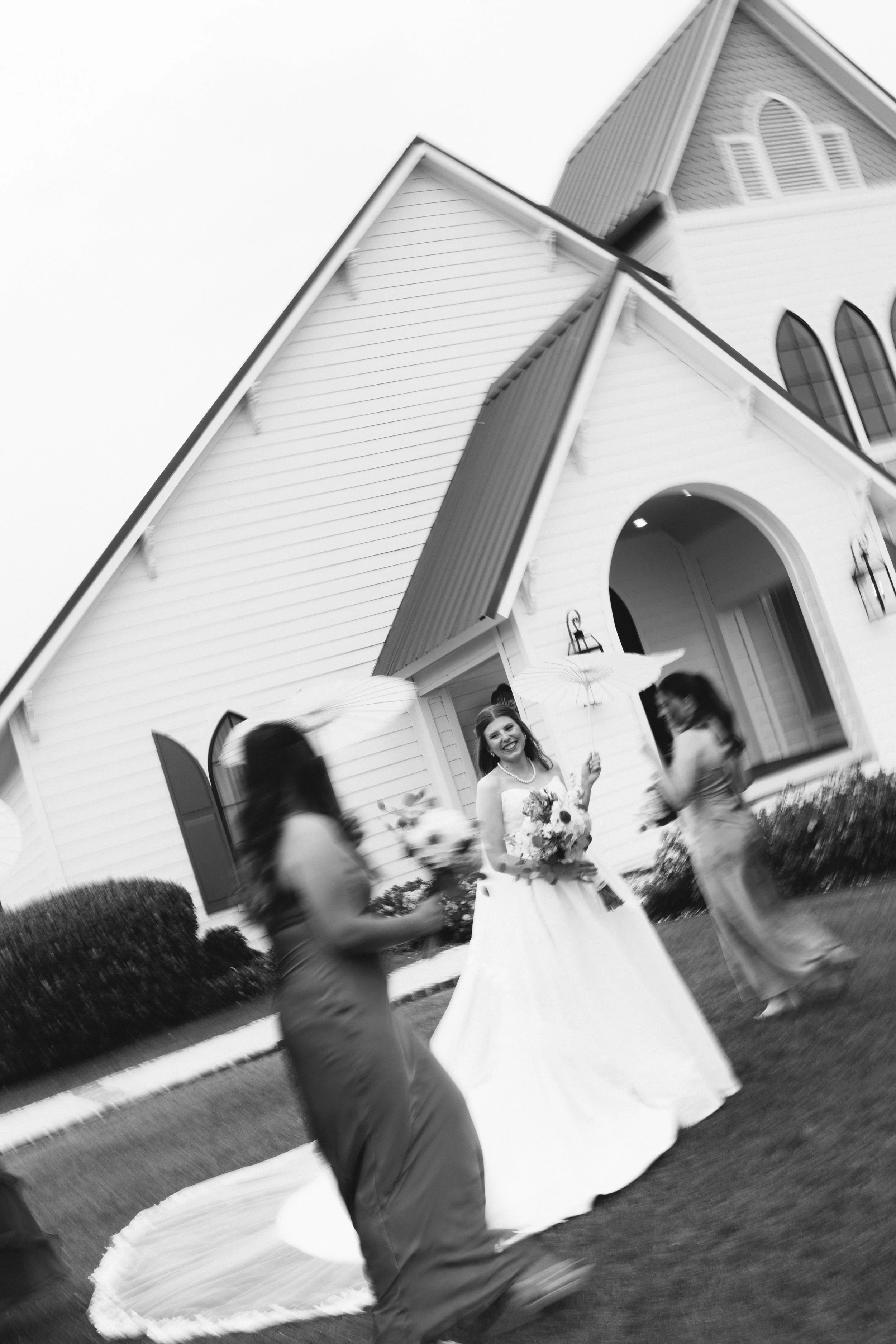 A bride in a wedding dress holding a bouquet, standing outside a church, smiling, with guests around her.