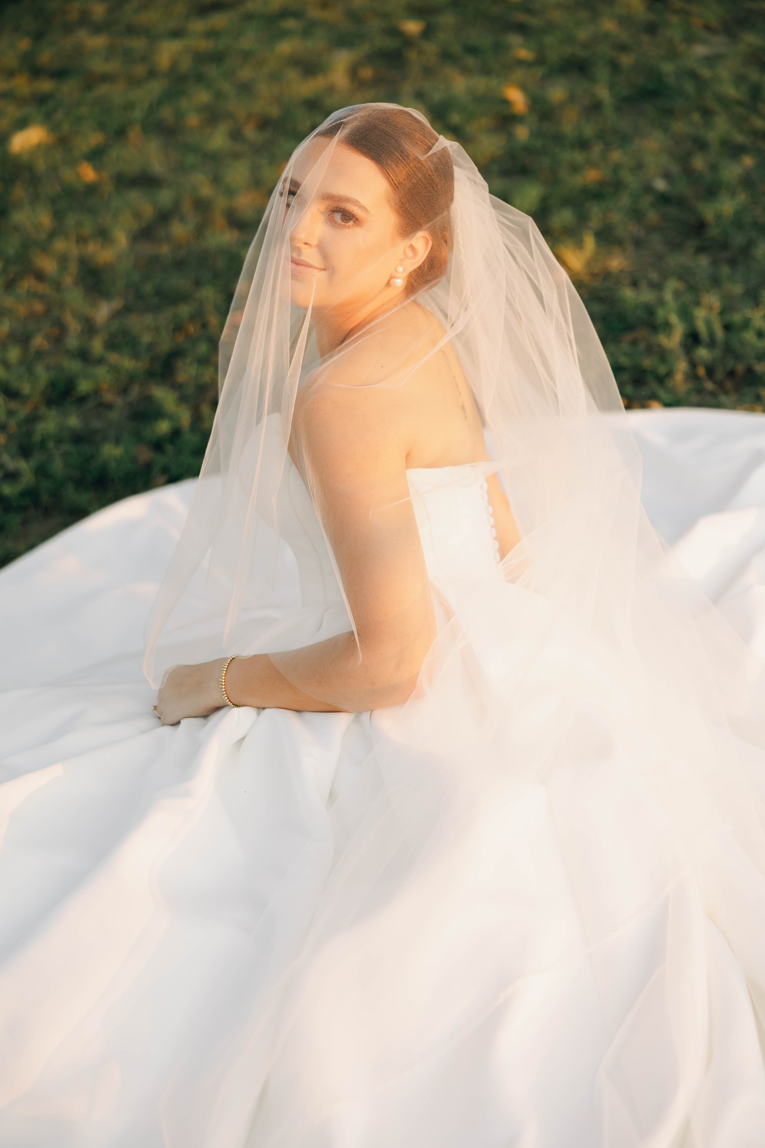 A bride sitting outdoors on a white wedding dress with a veil, looking over her shoulder at the camera.