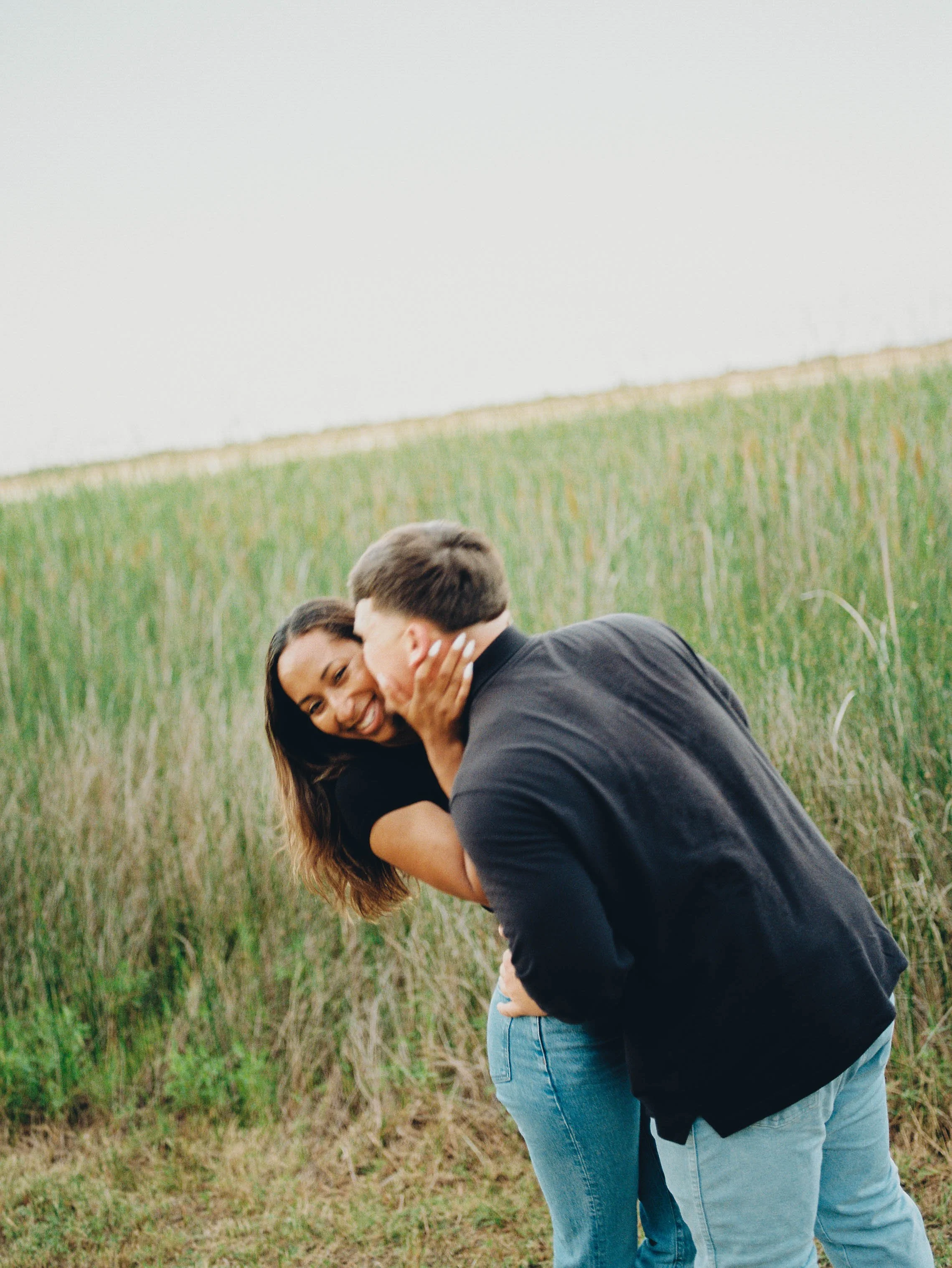 A couple, a woman and a man, sharing an intimate moment outdoors near a grassy field with a cloudy sky.