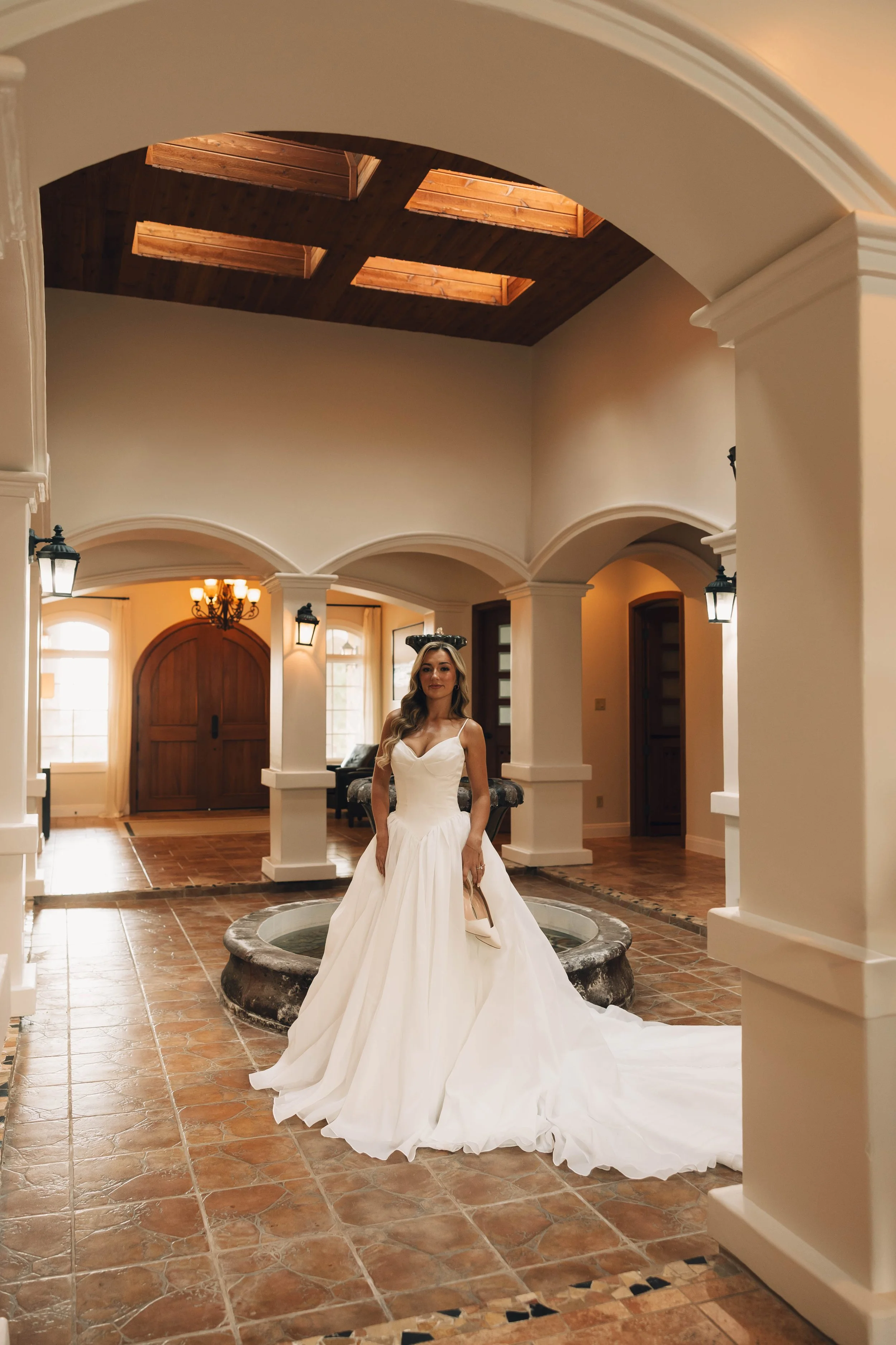 Woman in a wedding dress holding a high heel shoe inside a building with tile flooring, white walls, and arched doorways.