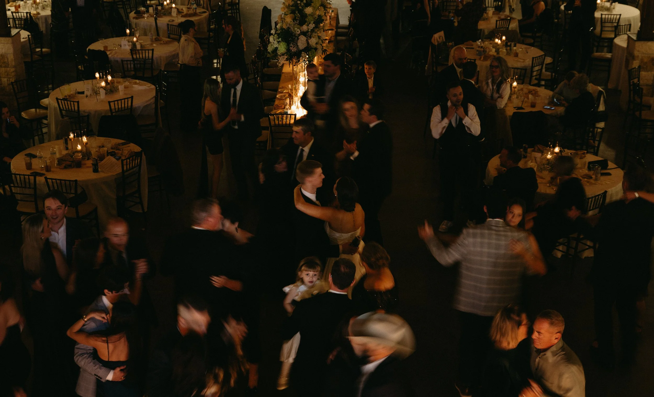 People dancing and socializing at an indoor wedding reception with decorated tables and dim lighting.