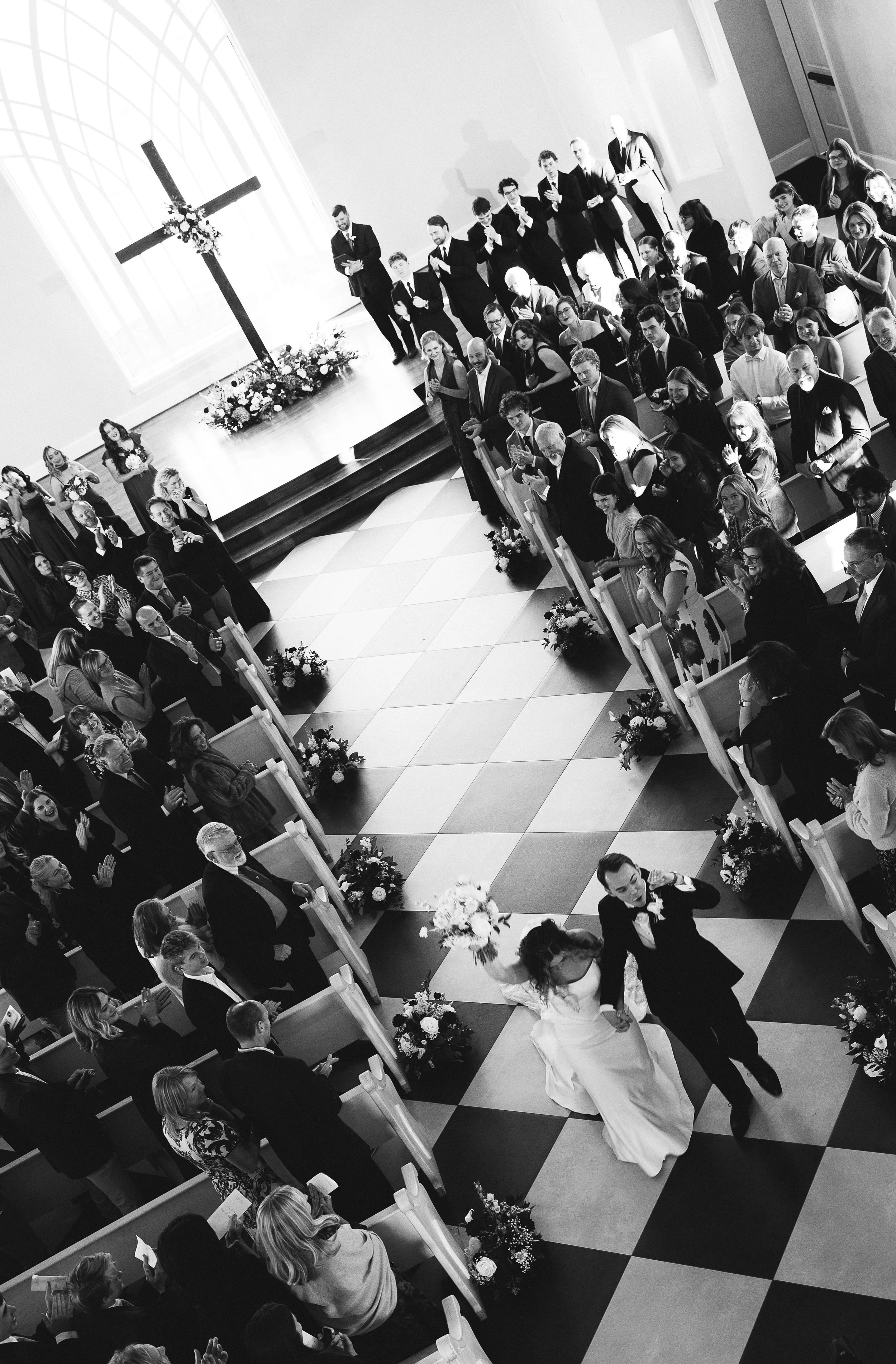 Black and white photo of a wedding ceremony in a church with guests standing and clapping as the bride and groom walk down the aisle.