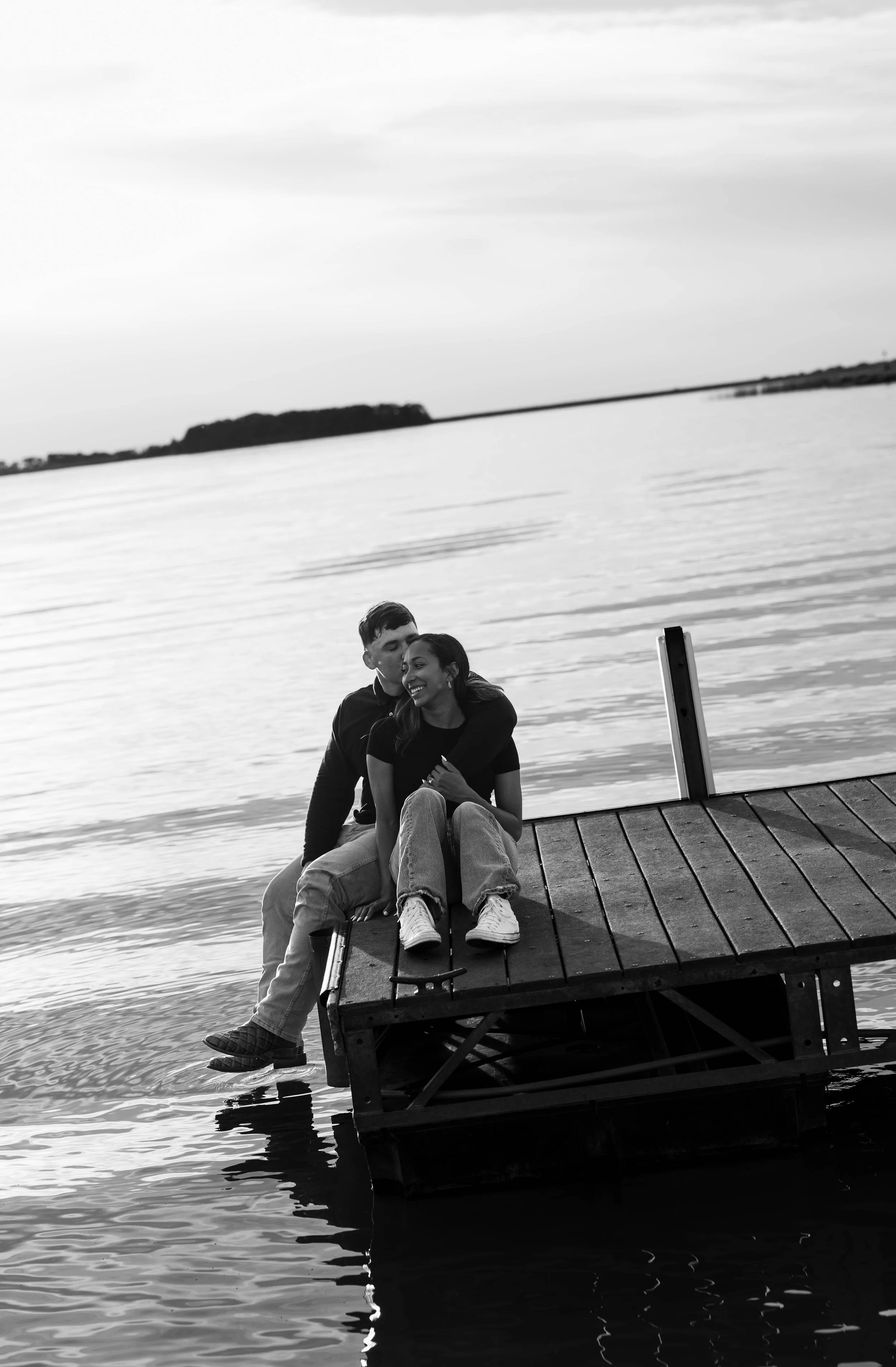 A black and white photo of a couple sitting on the edge of a dock by the water, embracing each other. One person is sitting while the other is leaning in close, with trees and a distant shoreline visible across the water.