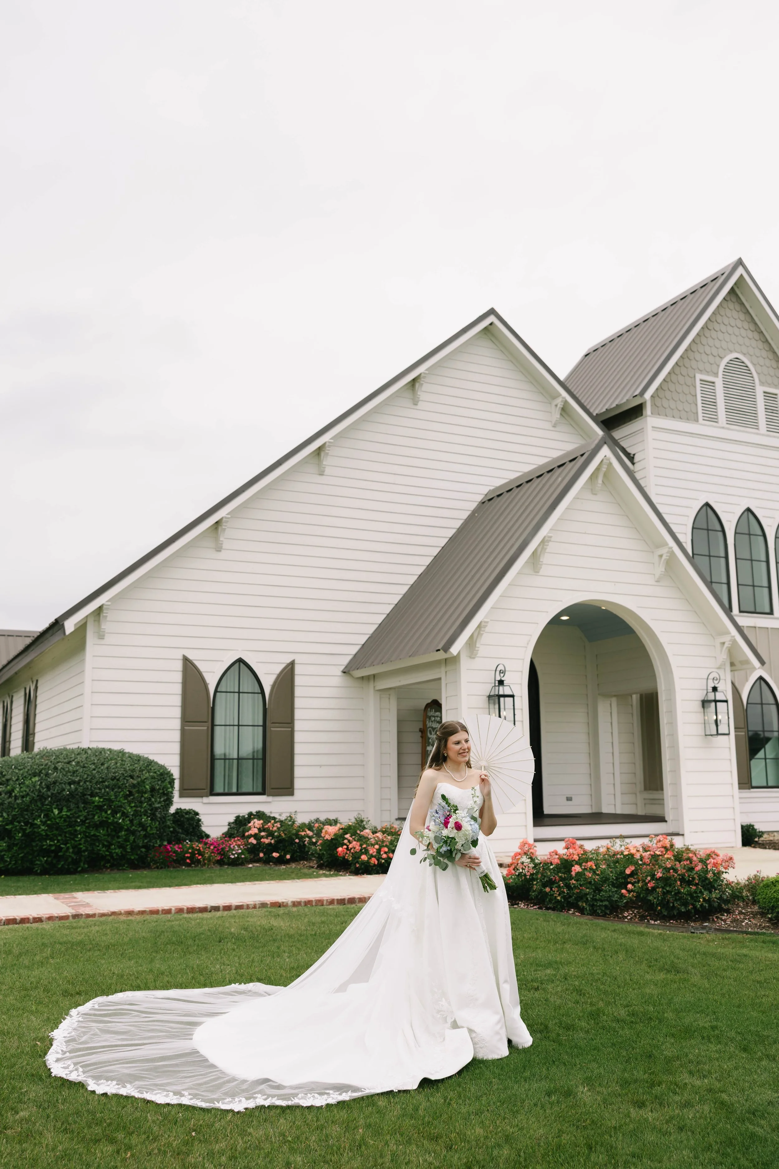 Bride in a white wedding gown holding a bouquet and a parasol, standing on a green lawn in front of a white church with dark brown shutters, surrounded by colorful flowers.