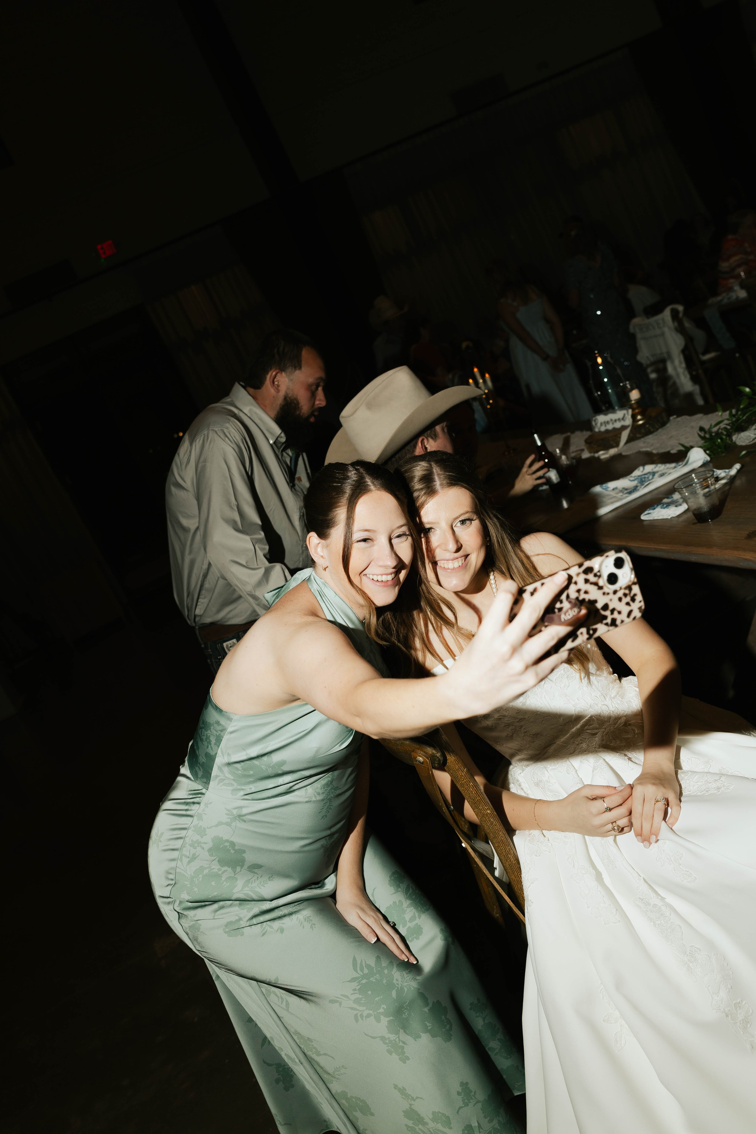 Two women taking a selfie at a formal event, with one in a light green dress and the other in a white dress, seated at a table. People are in the background standing and talking in a dimly lit room.