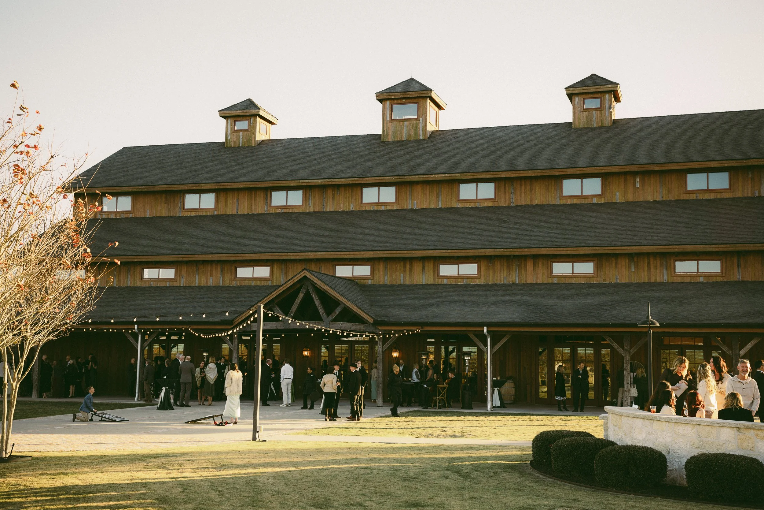 People gathering outside a large rustic wooden building with string lights, in a grassy area with shrubs and a leafless tree.
