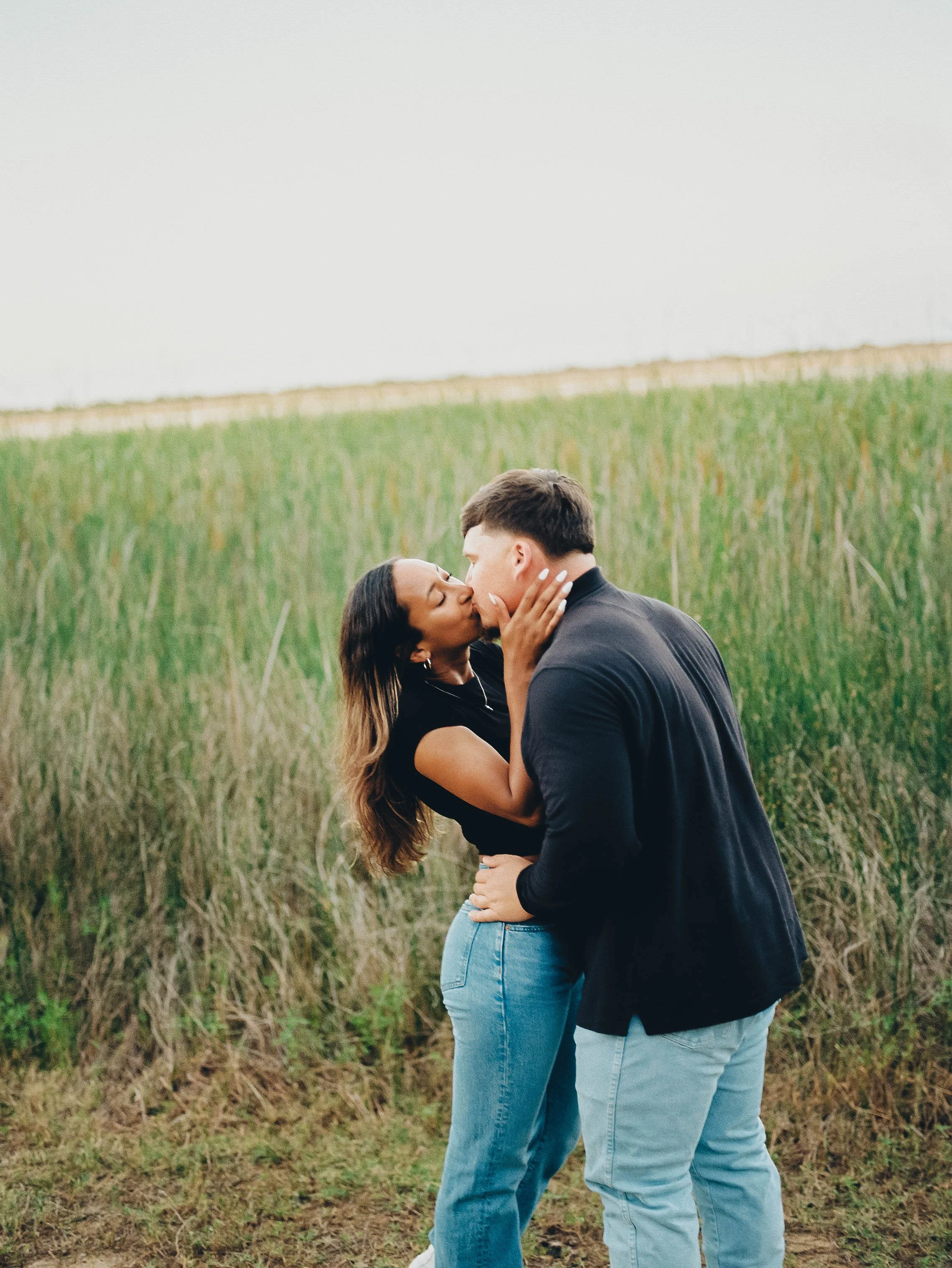 A couple standing close together in a grassy field, gazing into each other's eyes, with a woman touching the man's face and both smiling.