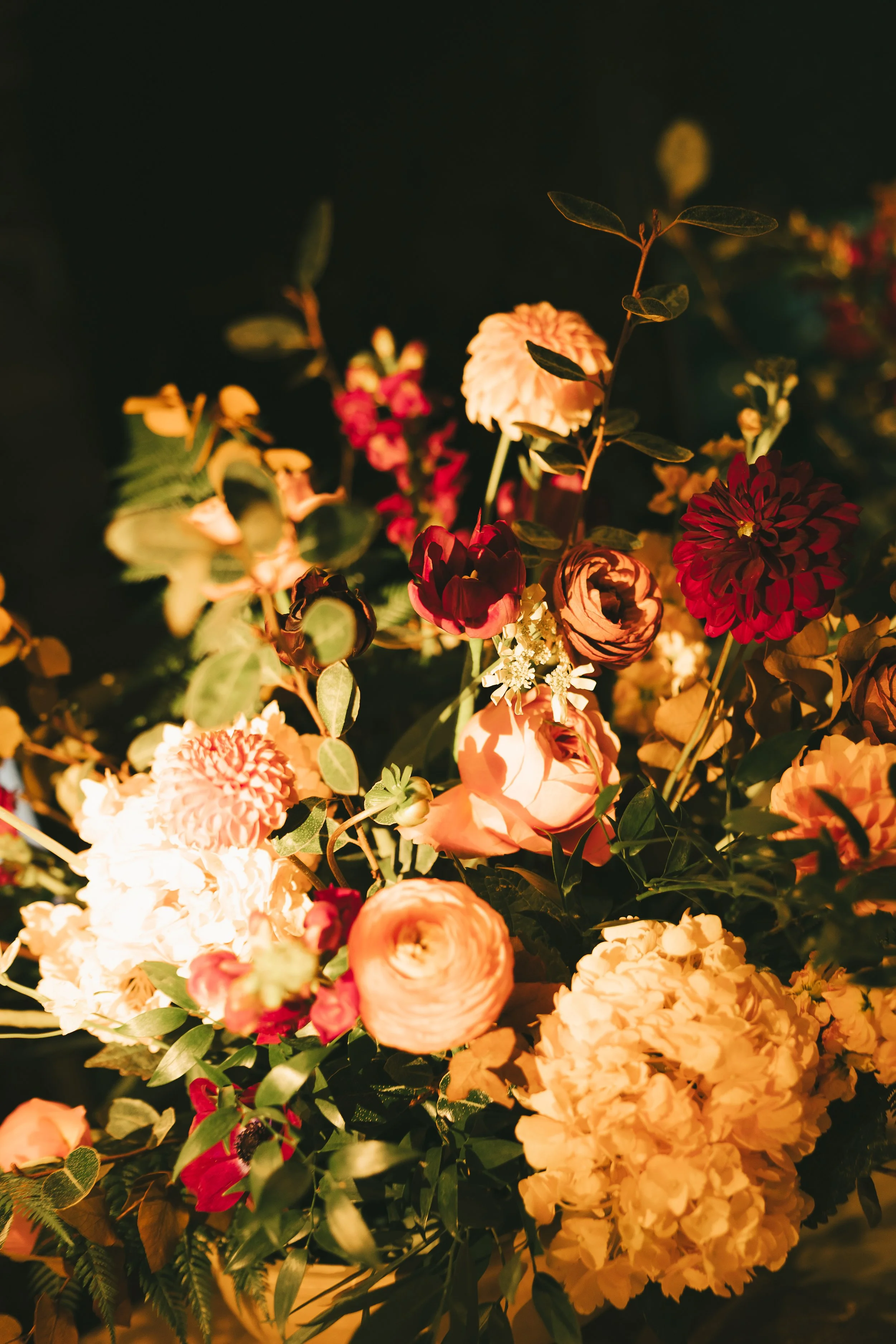 A close-up of a vibrant bouquet of mixed flowers, including roses, dahlias, and ranunculus, with dark background lighting.
