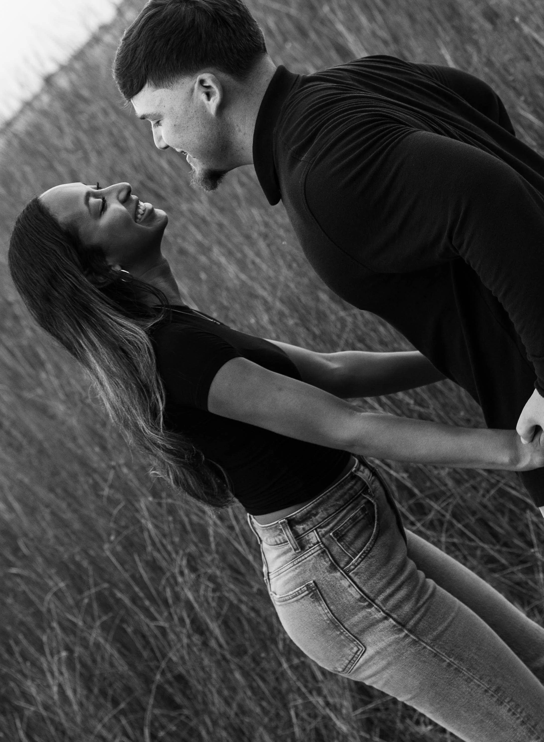 A black and white photo of a couple in a field, facing each other, smiling and leaning in close, with the woman wearing a black top and high-waisted jeans, and the man in a dark shirt and jeans.