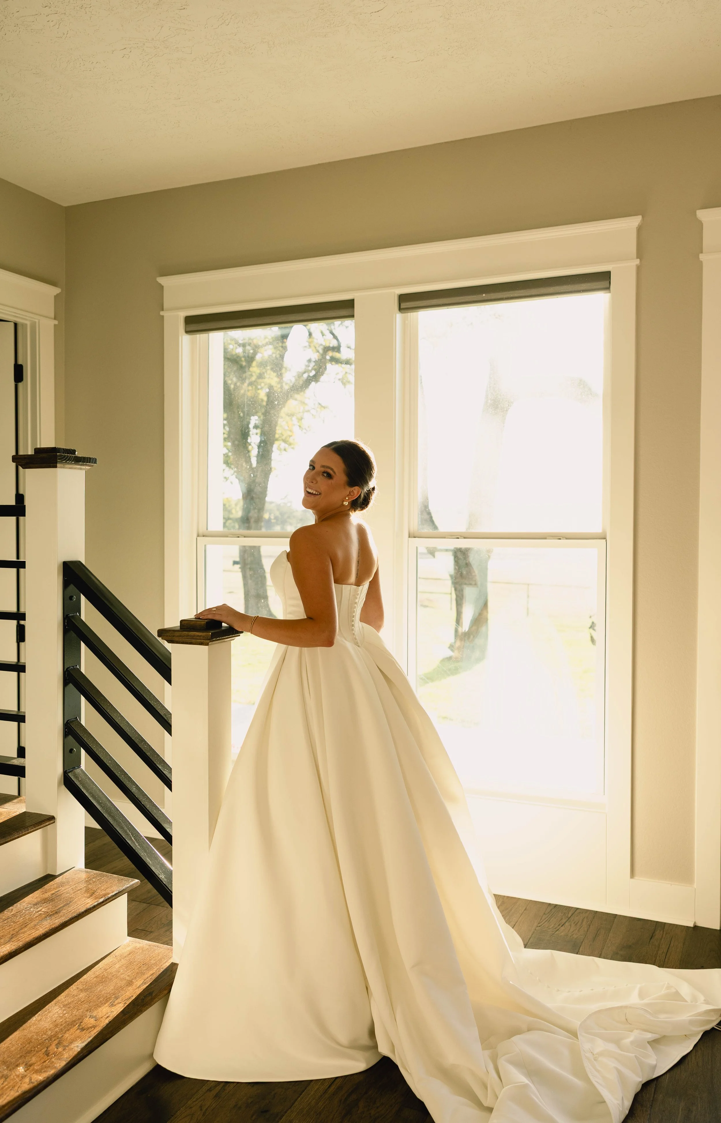 A bride in a strapless white wedding gown smiling and posing near a window in a well-lit room.