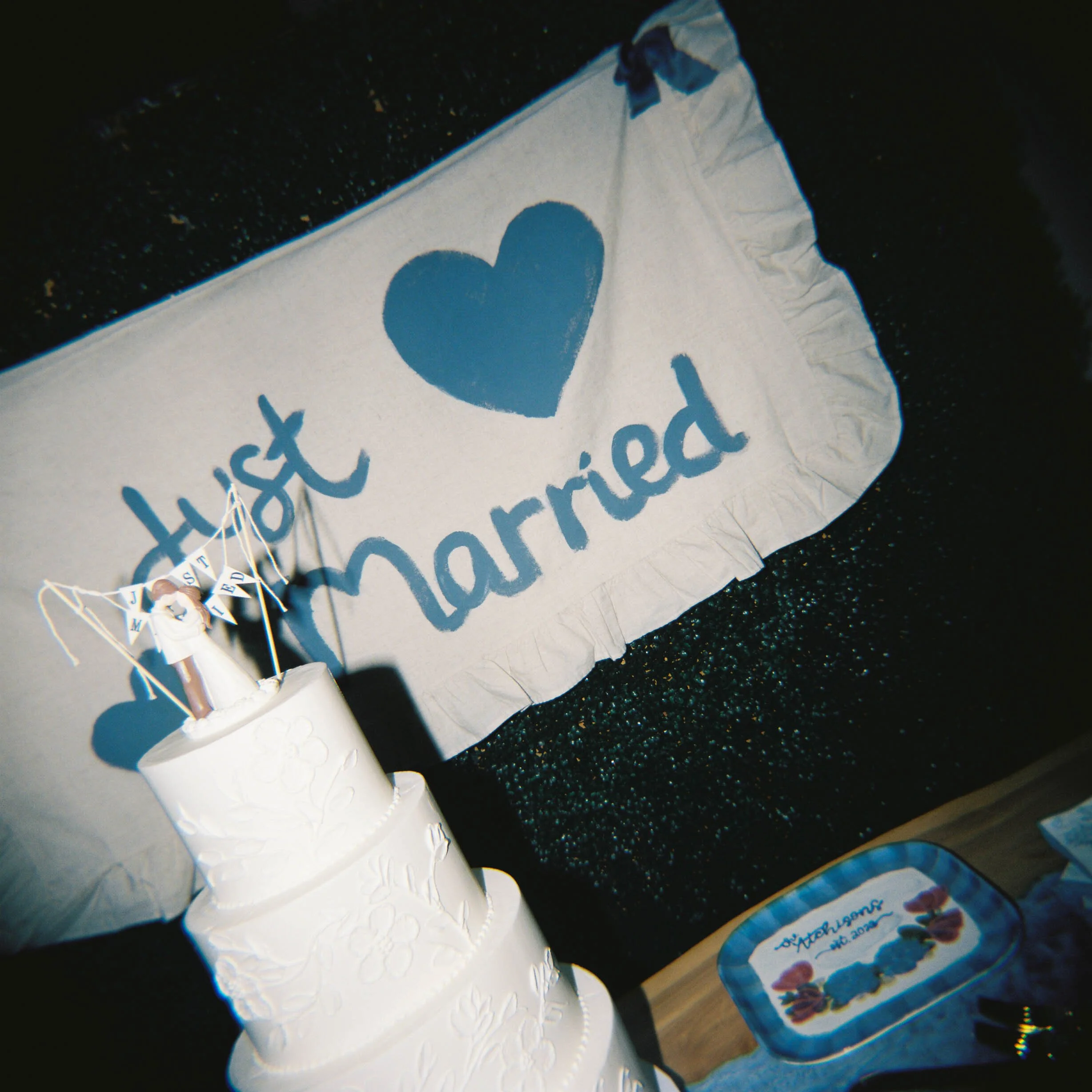 A birthday cake with white icing and embossed heart patterns, decorated with a small figurine of an angel holding balloons. Part of a cake box with colorful lettering is visible at the bottom of the image.