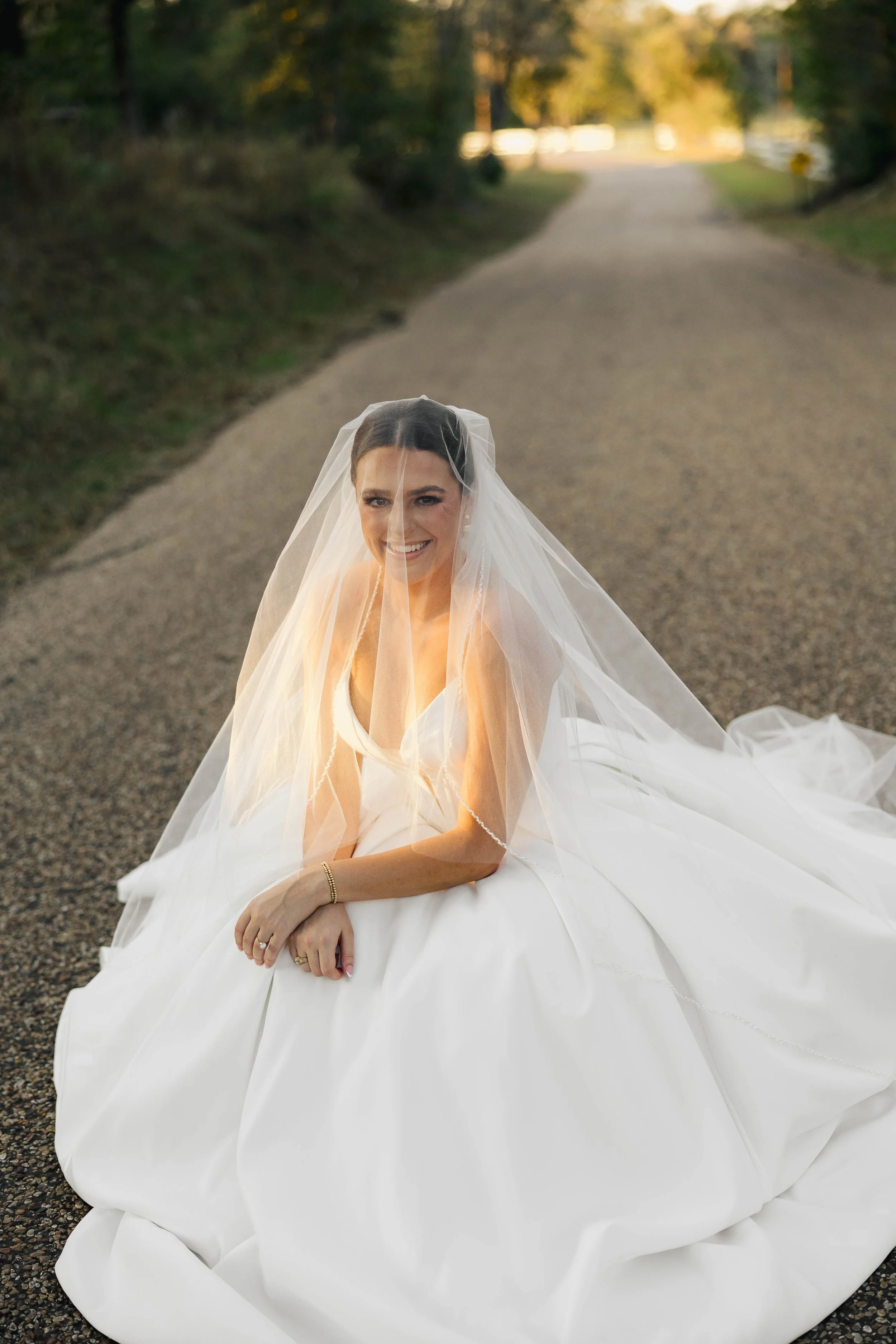 A bride in a white wedding gown and veil sitting on a rural road at sunset, smiling at the camera.